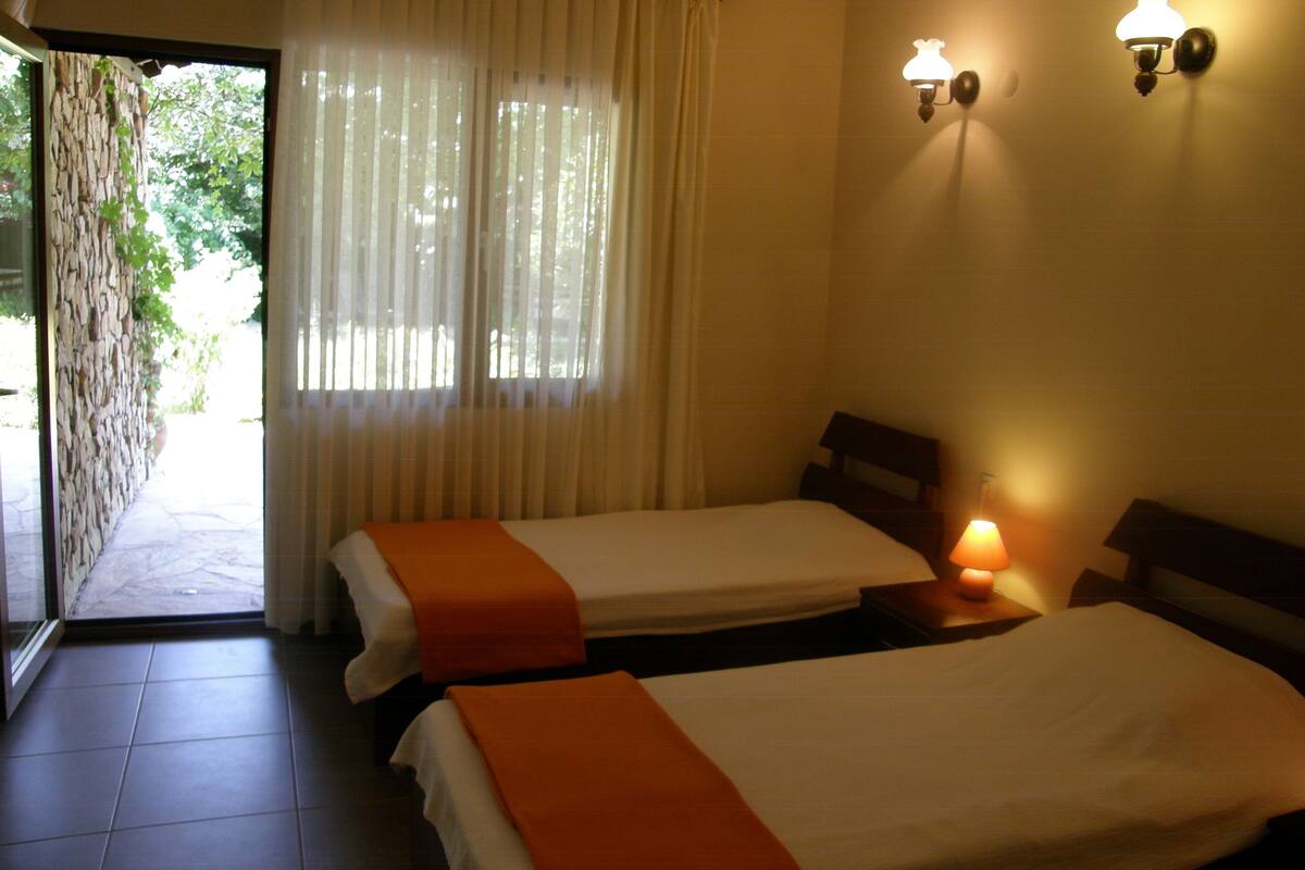 A hotel room with two beds, a window with curtains, a stone wall, and a nightstand with a lamp.