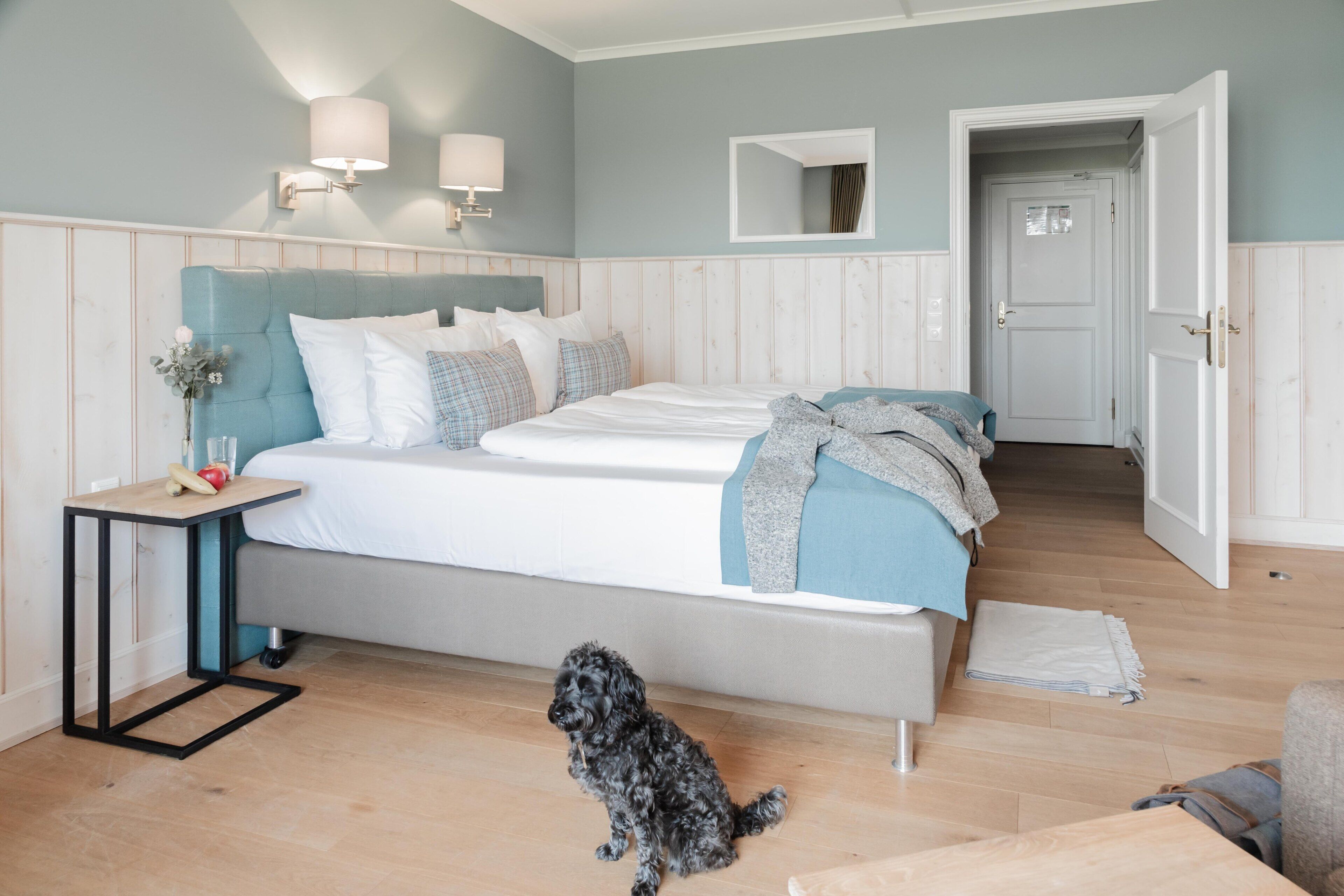 Et soveværelse med seng, natbord, lampe, spejl og en hund.