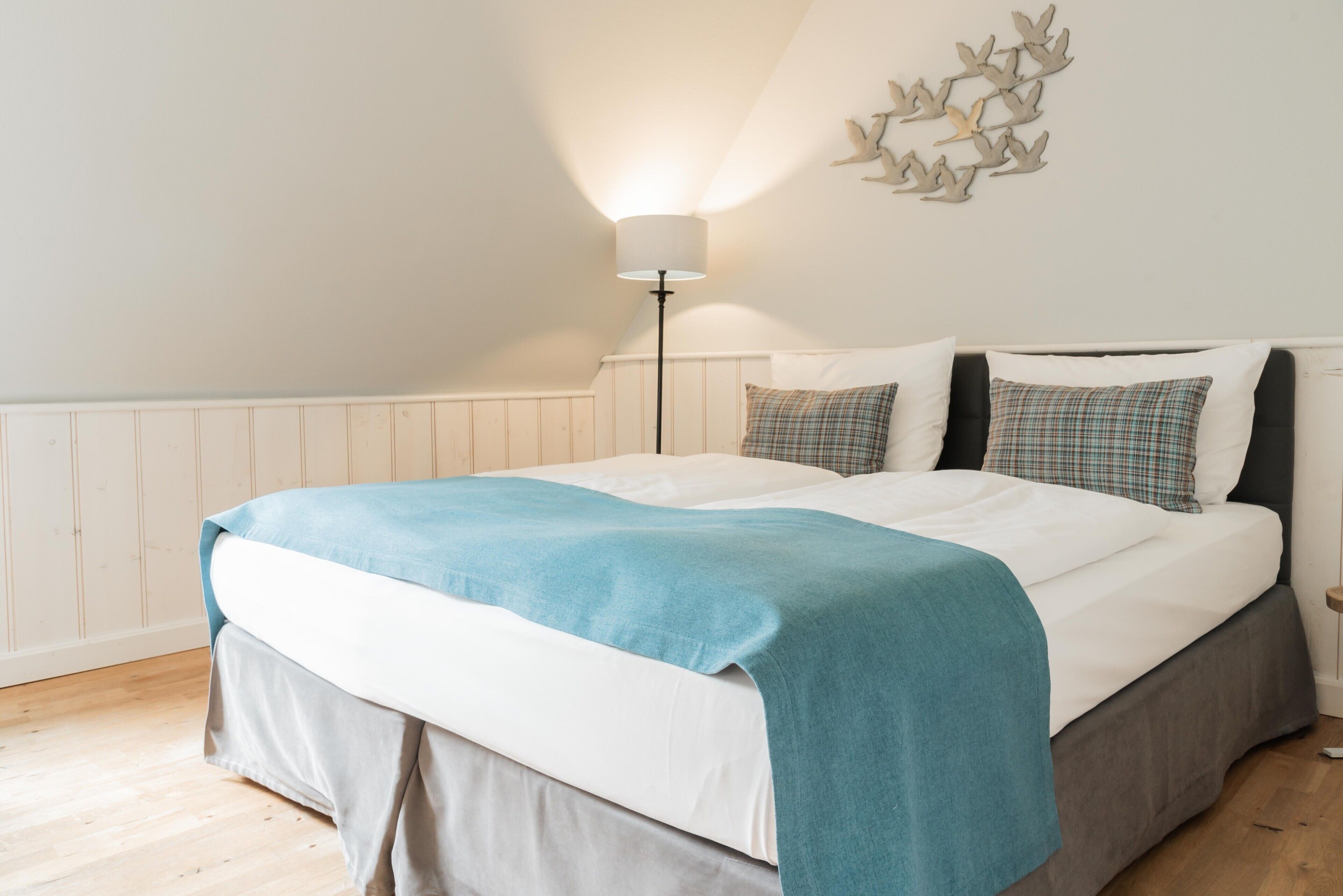A neatly made bed with a blue blanket, white pillows, and a grey headboard. A floor lamp with a white shade is positioned to the left of the bed.