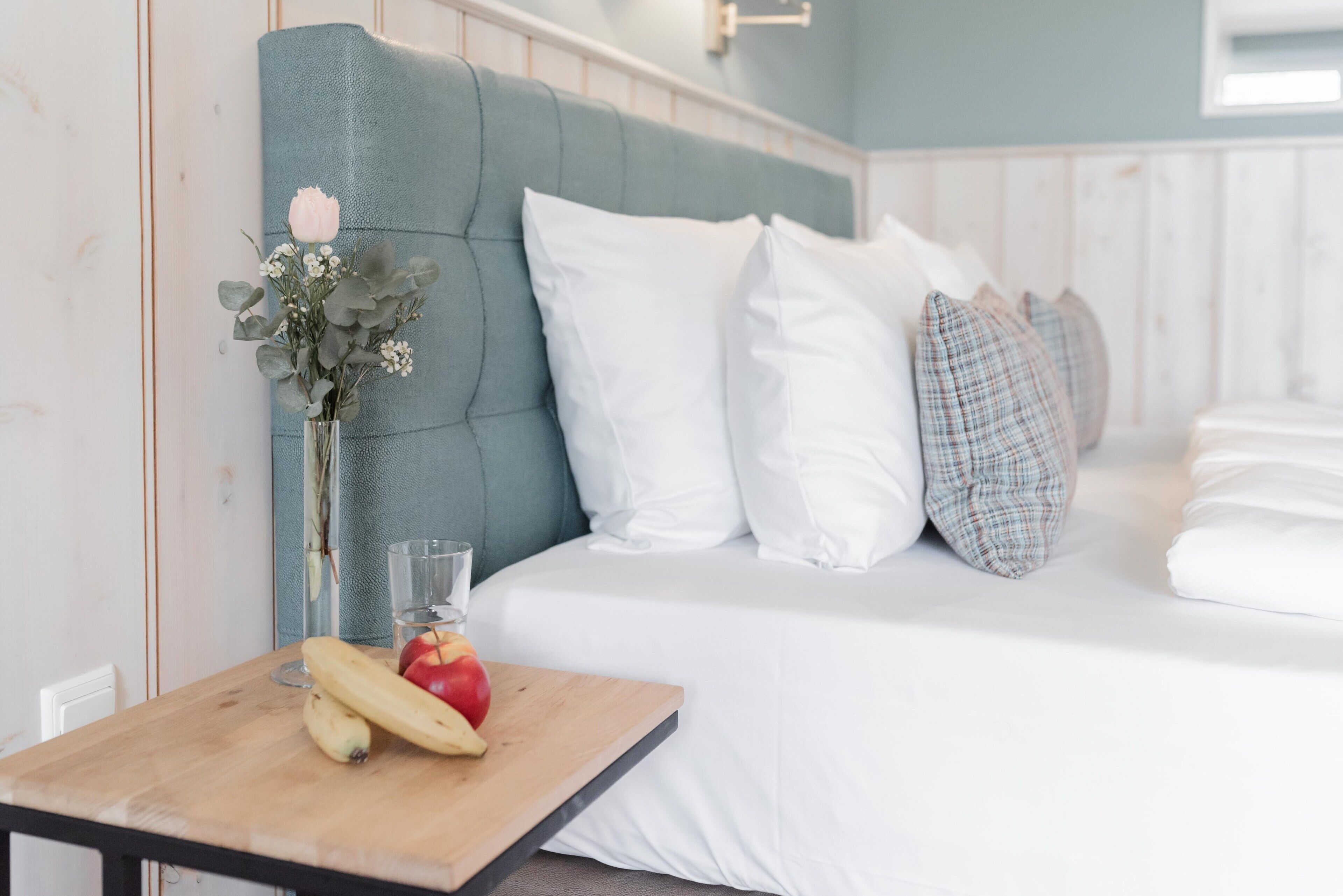 A neatly made bed with white bedding and a teal headboard, a small wooden table with a glass of water and fruit, and a vase with flowers.