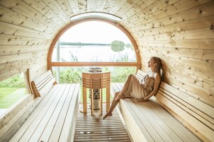 Eine Holzhütte mit einem großen Rundfenster mit Blick auf einen See. Im Inneren befindet sich eine hölzerne Bank und ein kleiner Tisch mit einem Wasserkocher.