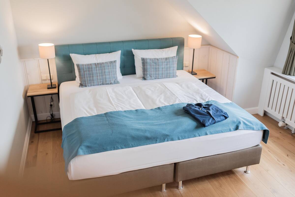 Ein ordentlich bezogenes Bett mit blauer und weißer Bettwäsche, zwei Nachttischlampen und einem Holznachttisch.