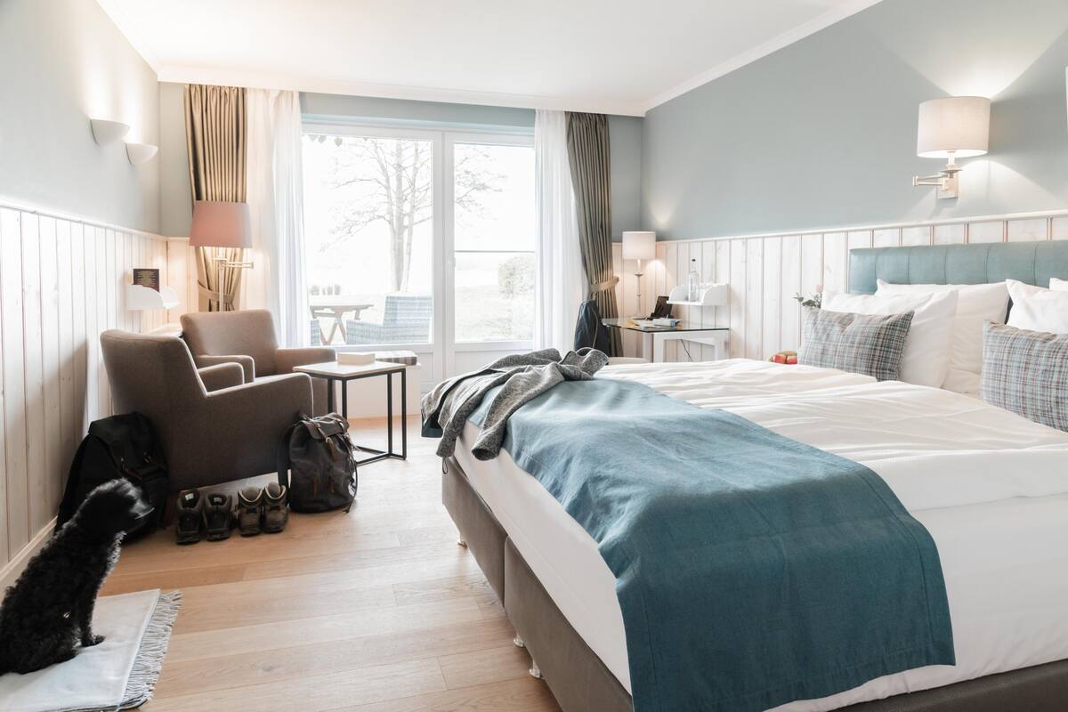 Ein Hotelzimmer mit Bett, Sessel, Tisch, Lampe und Fenster.