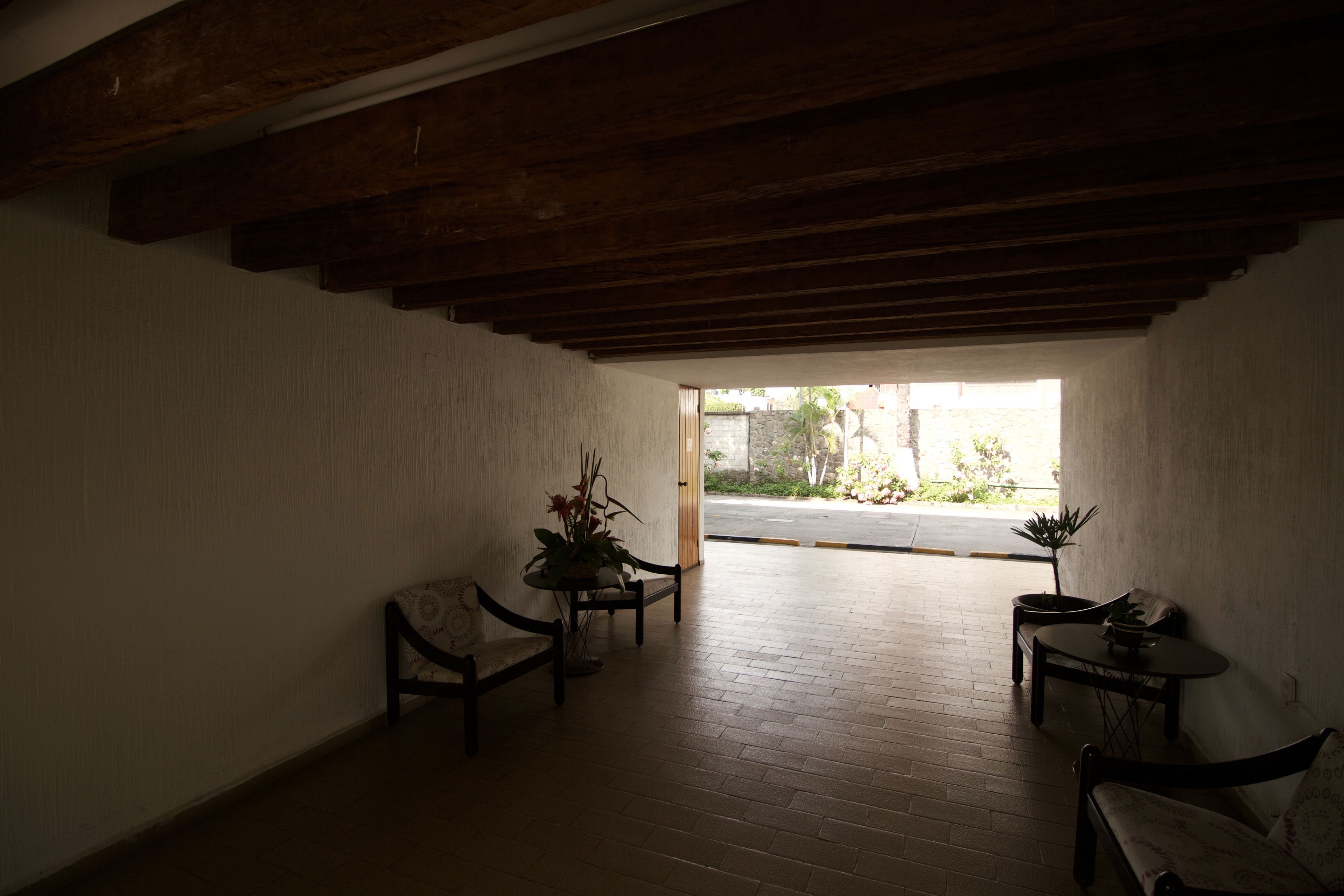 Un espacio interior con techo de madera, suelo de ladrillo y vistas al exterior a través de un ventanal abierto. Hay dos mesas con sillas y una planta en maceta.