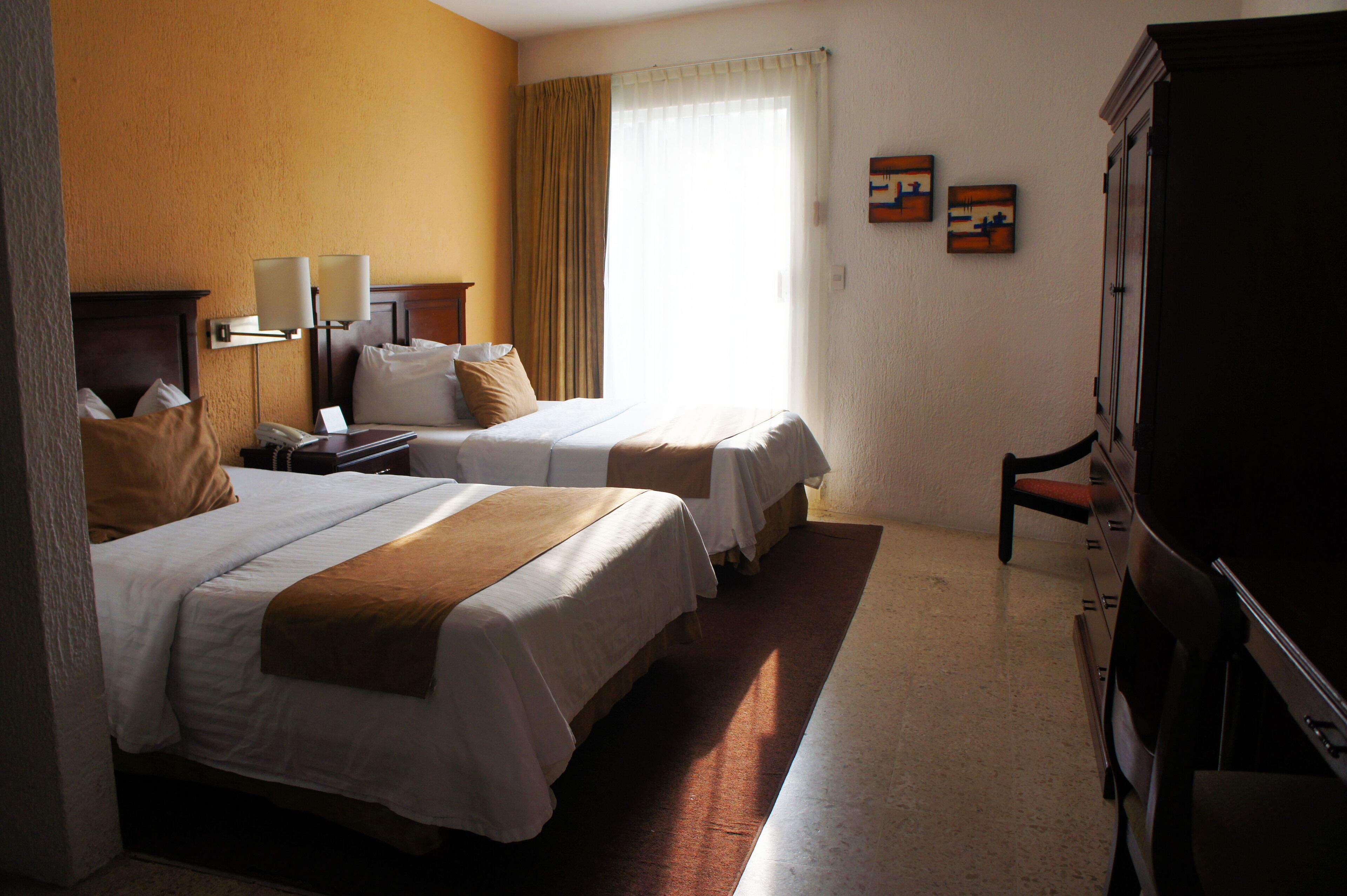 Una camera d'albergo con due letti, una scrivania, una sedia, una televisione e opere d'arte alle pareti.