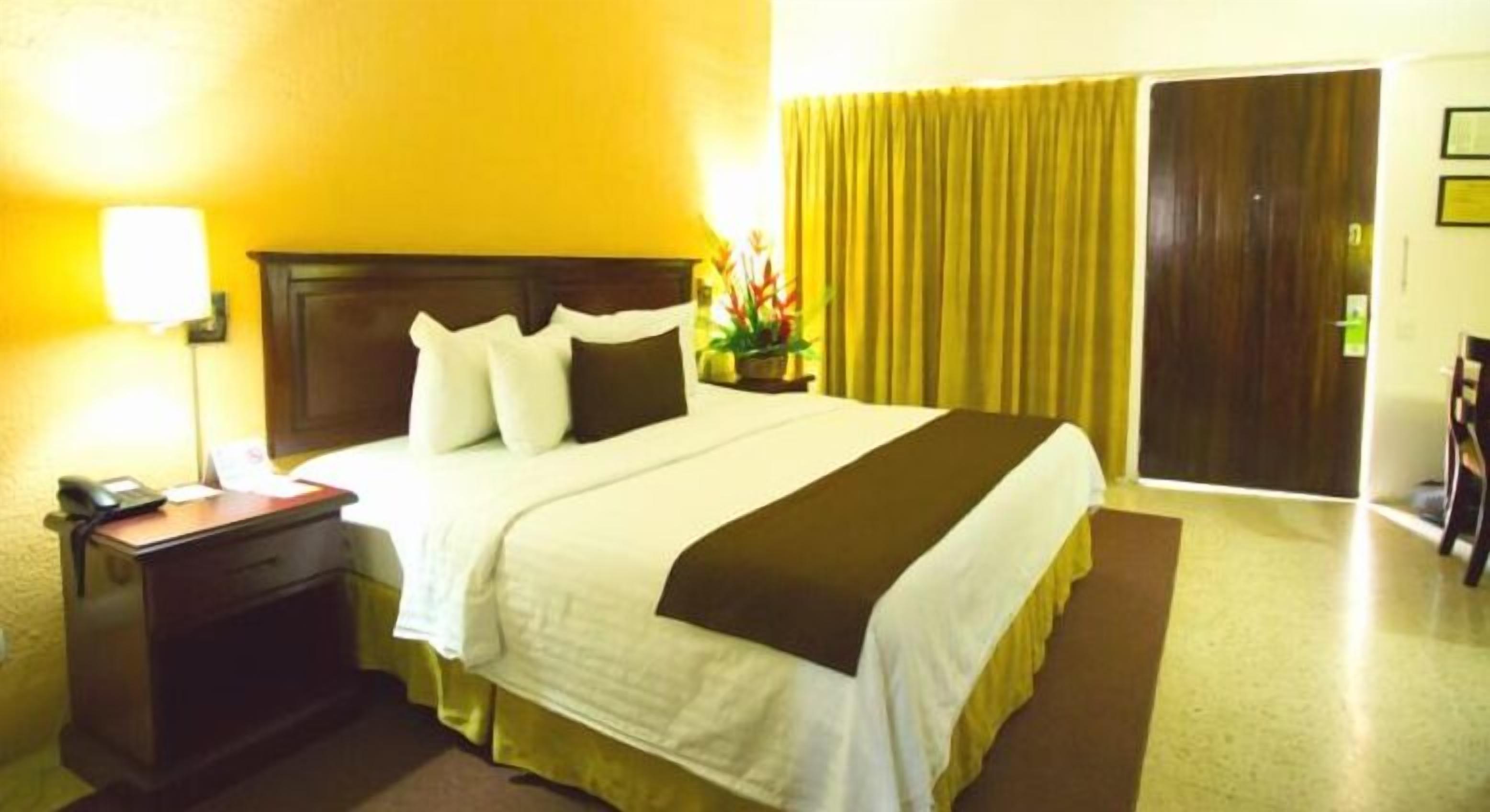 Habitación de hotel con cama, escritorio con teléfono, mesita de noche, lámpara y ventana con cortinas.