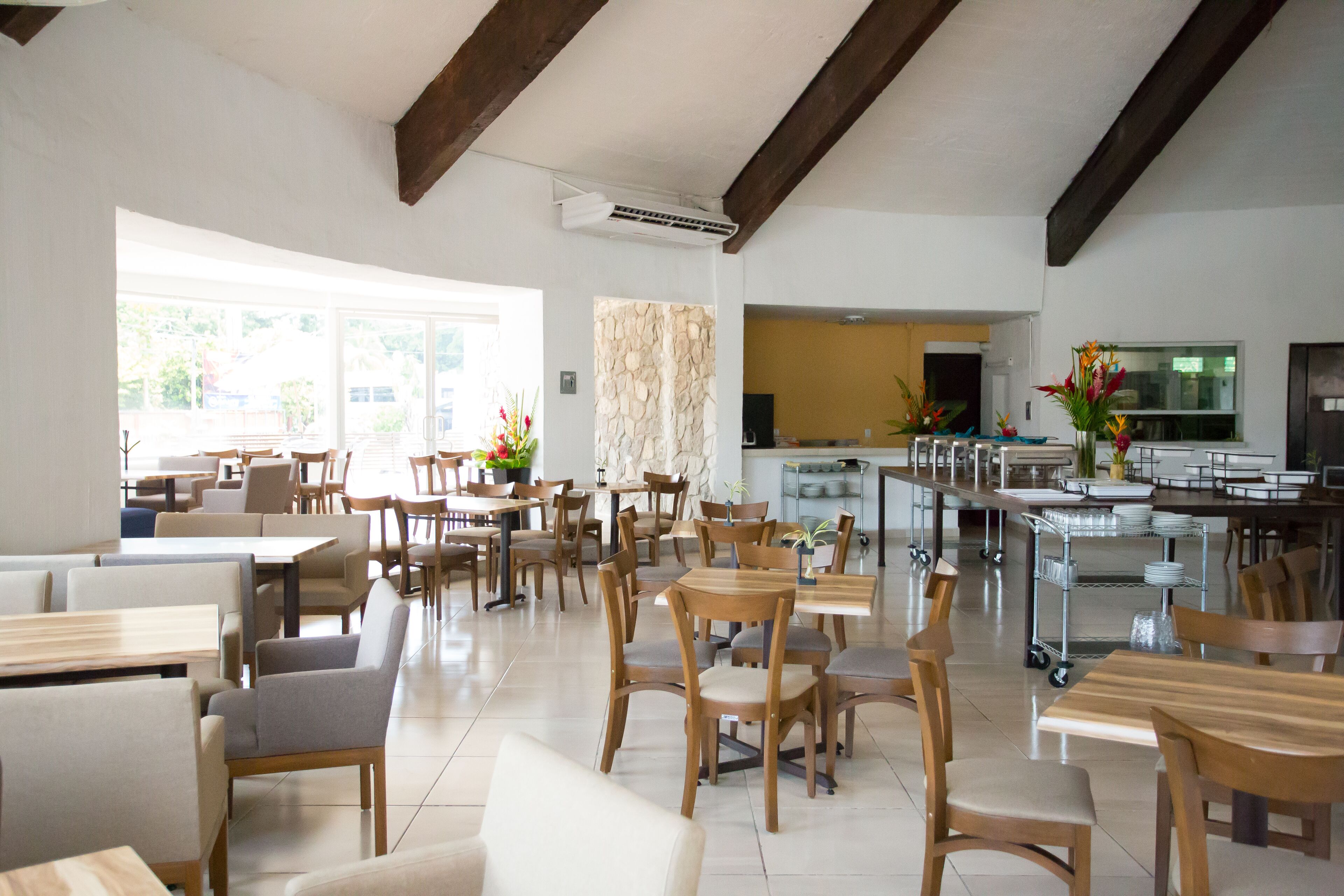 Un vaste espace de salle à manger avec des tables et des chaises en bois, une cuisine à l’arrière-plan et de grandes fenêtres qui laissent entrer la lumière naturelle.