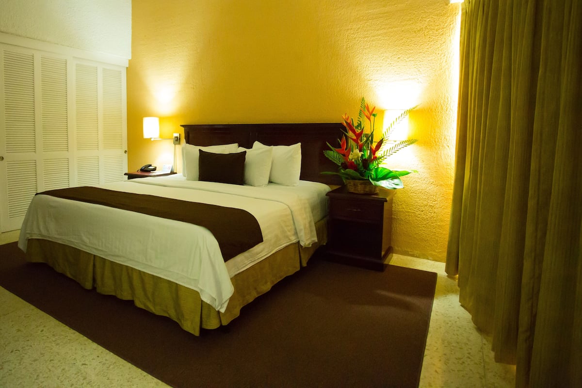 A hotel room with a large bed, a nightstand with a lamp, and a door with louvered shutters.