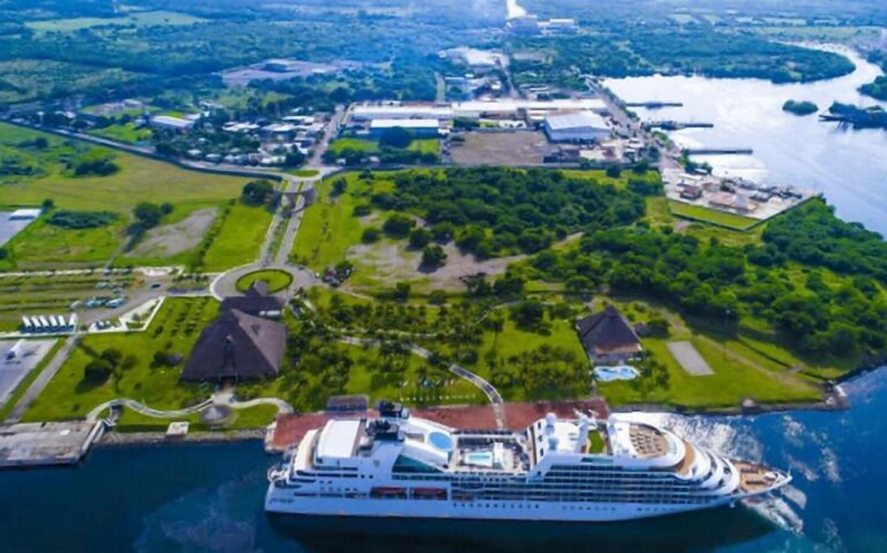 A large cruise ship docked at a port with a landscaped area and buildings in the background.