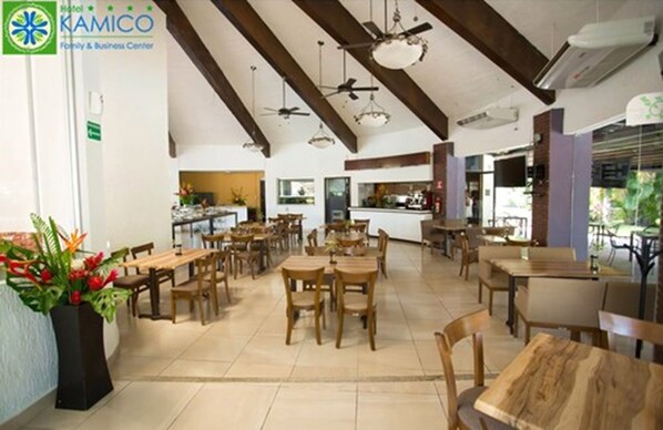 A hotel dining area with wooden tables and chairs, ceiling fans, and a kitchen in the background.