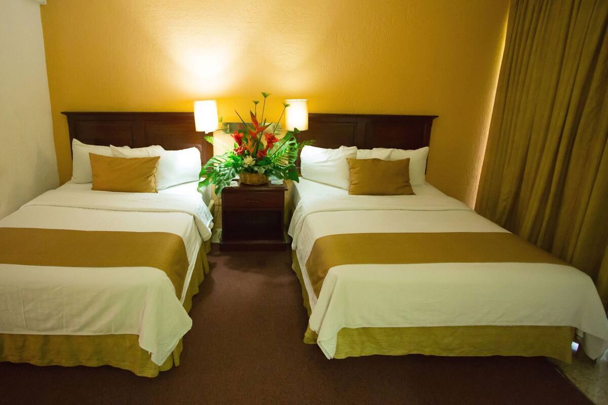 A hotel room with two beds, a nightstand with a lamp, and a floral arrangement.