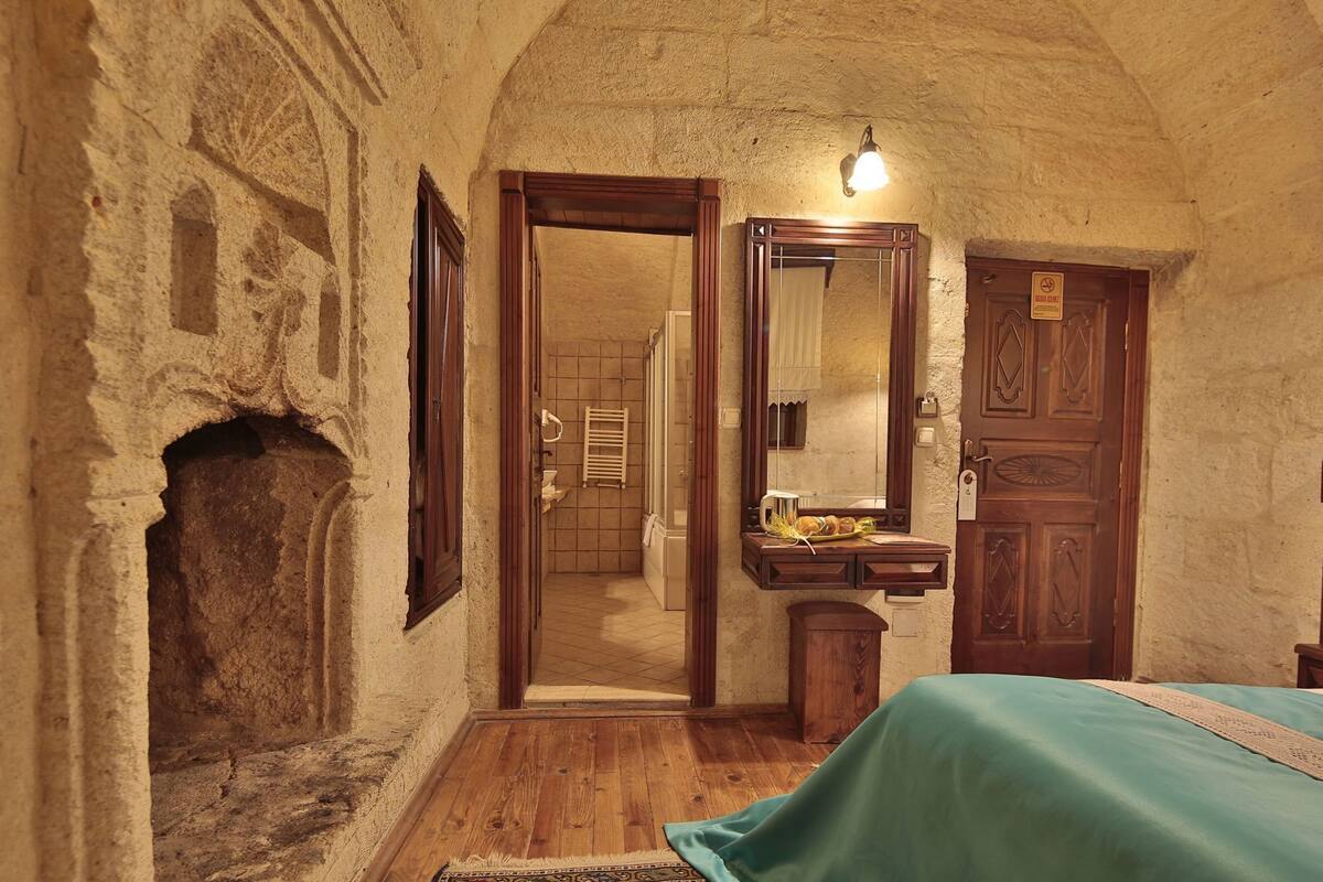 Ein Zimmer mit Steinwand, Holztür, einem Spiegel, einem kleinen Holztisch mit Handtuch und einem Bett mit grüner Bettdecke.