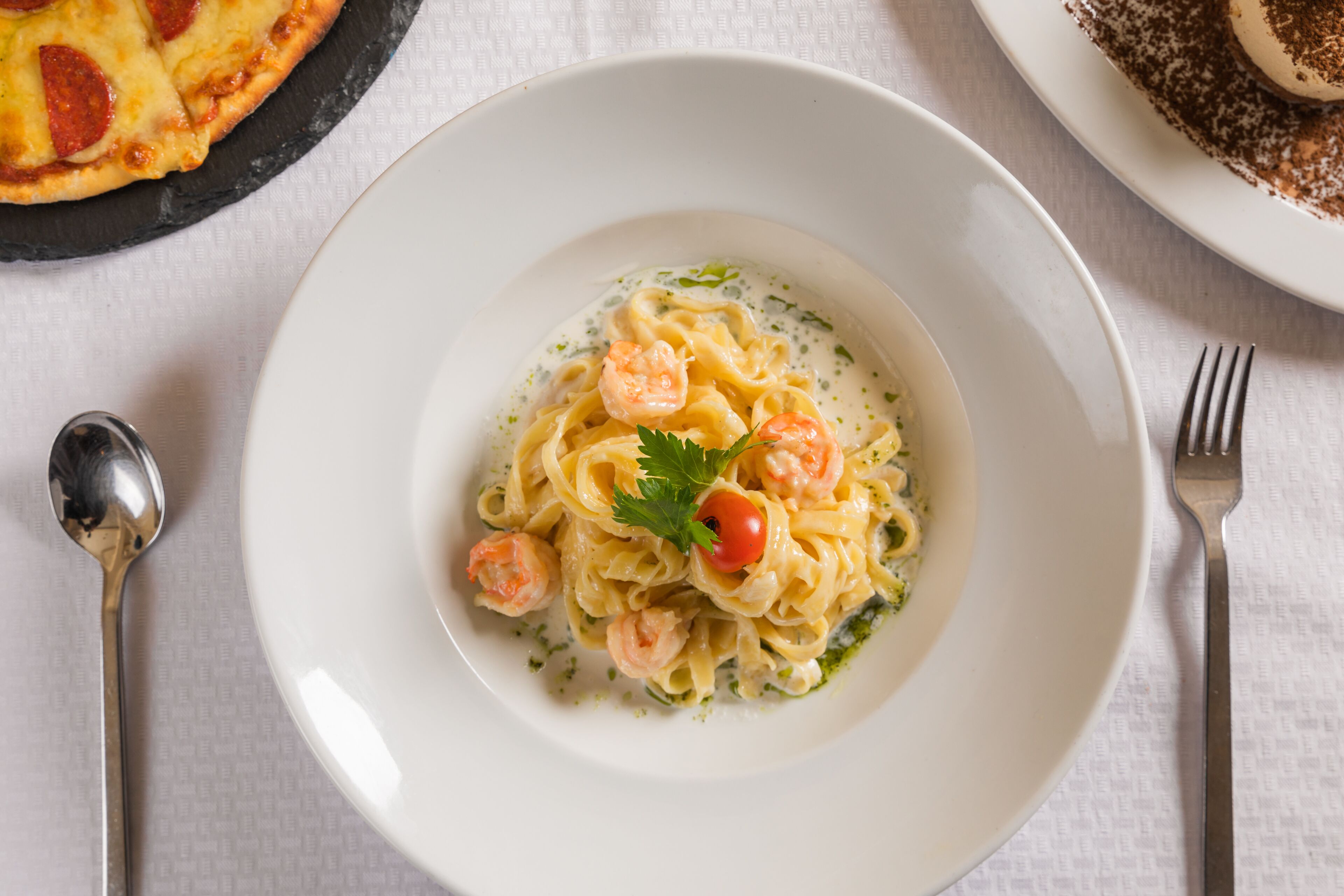 Un plato de pasta con camarones y una guarnición, acompañado de un tenedor y una cuchara sobre un mantel.