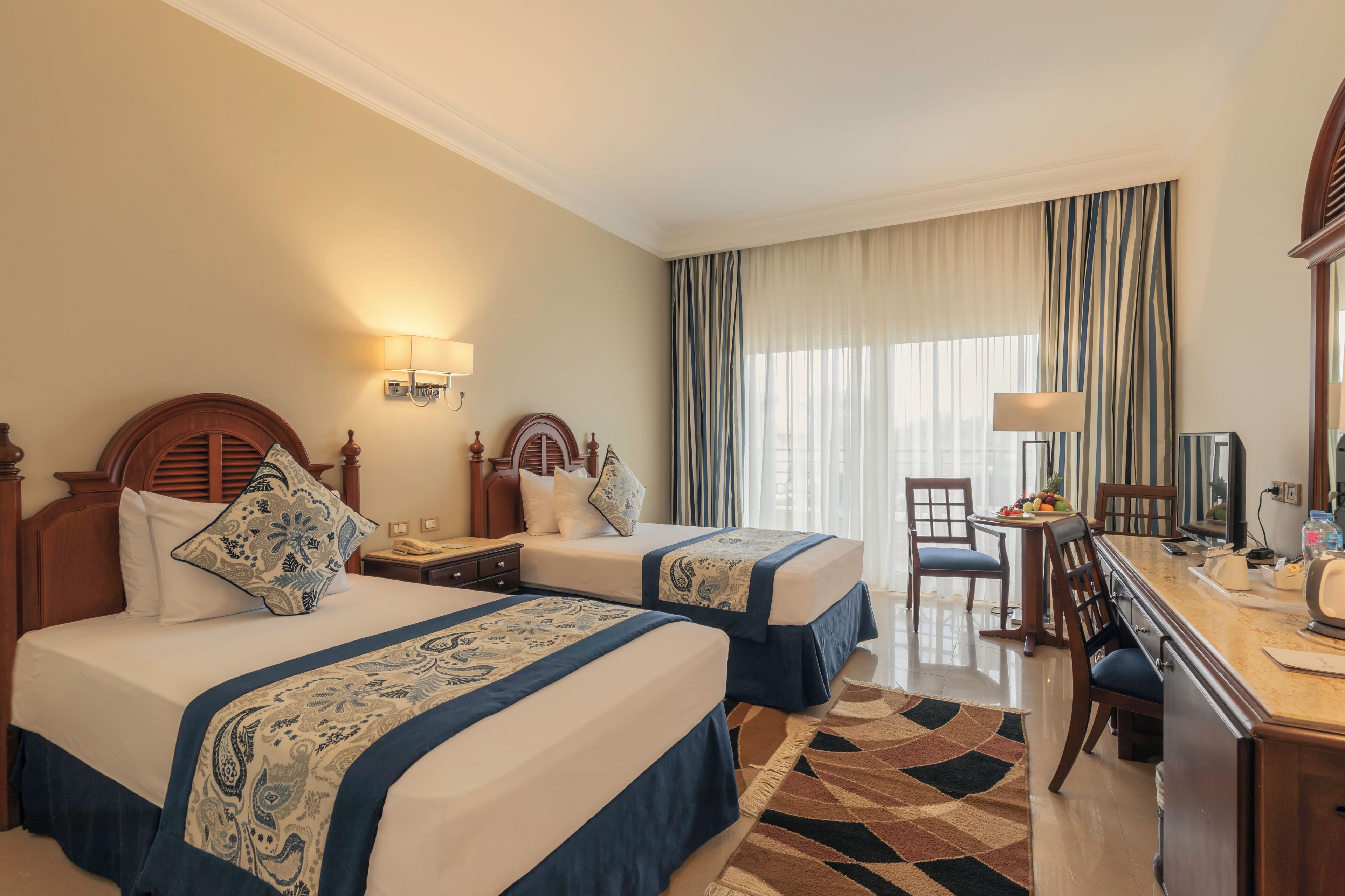 A hotel room with two beds, a desk, a chair, and a large window with curtains.