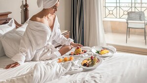 Een hotelkamer met een bed, een ontbijt aan een eettafel, gordijnen voor het raam en uitzicht op zee.