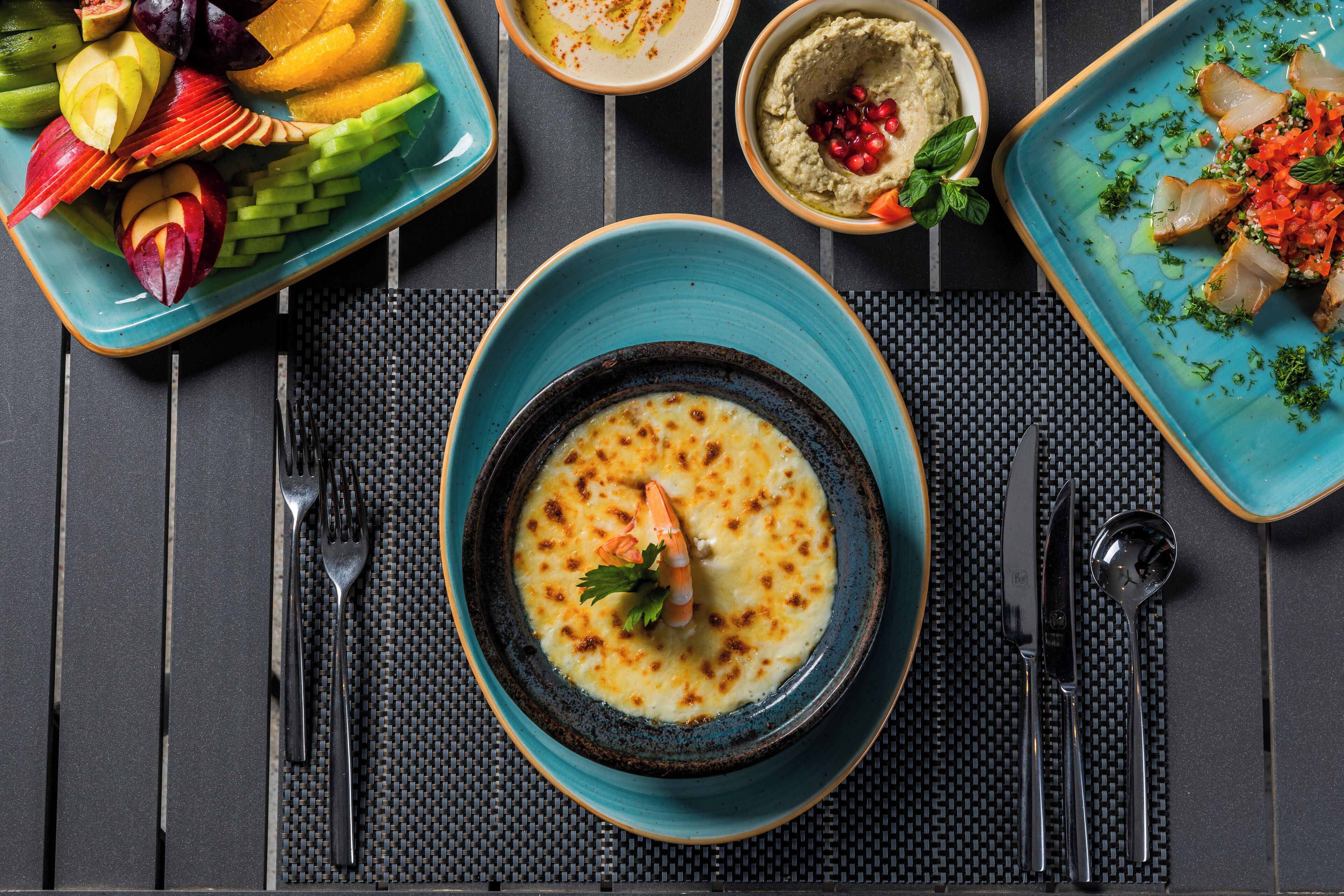 A dining table with a variety of dishes, including a bowl of soup with a shrimp garnish, a plate of sliced fruit, and a bowl of hummus with pomegranate seeds.