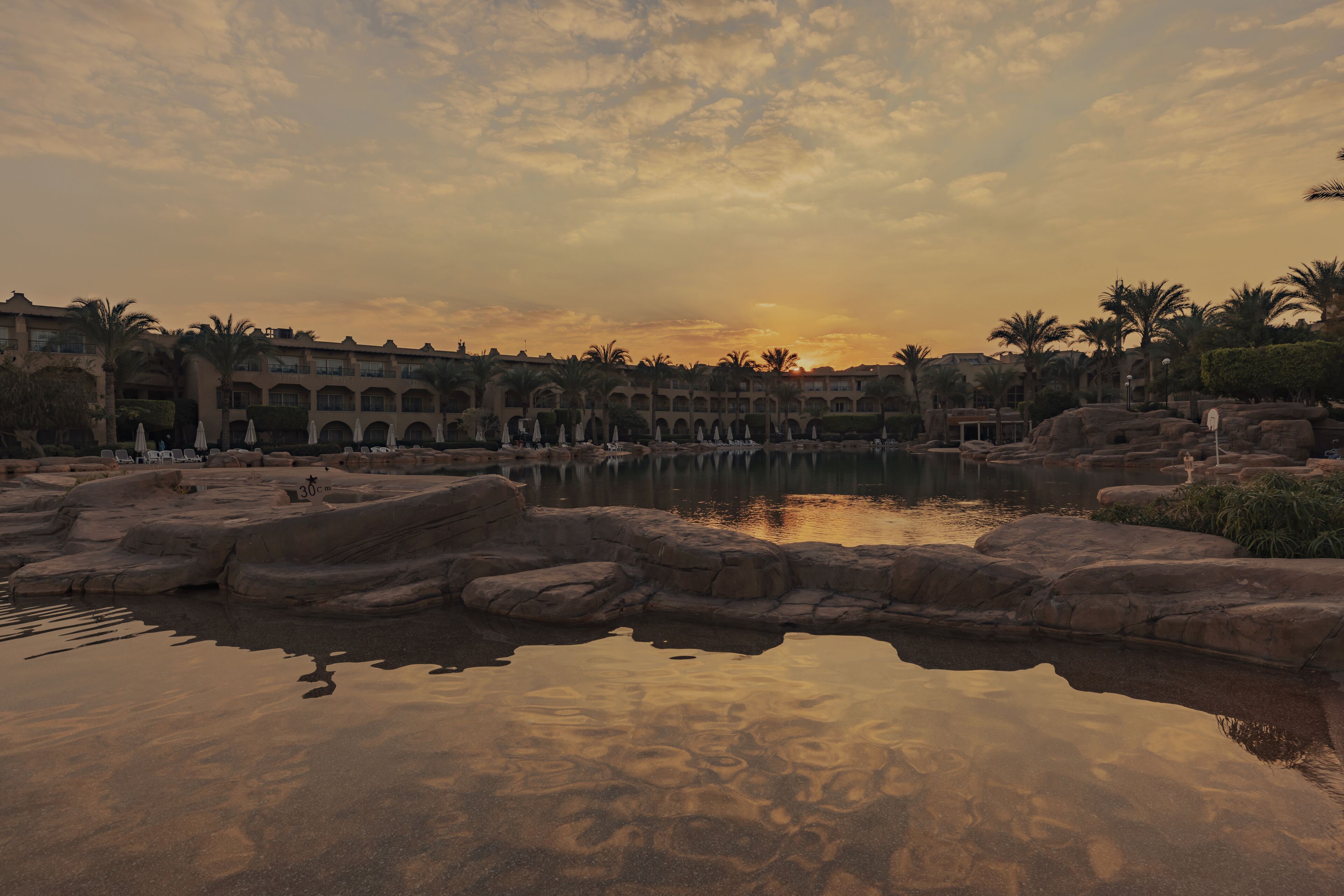 A resort with a pool surrounded by rock formations and palm trees, reflecting the sunset.