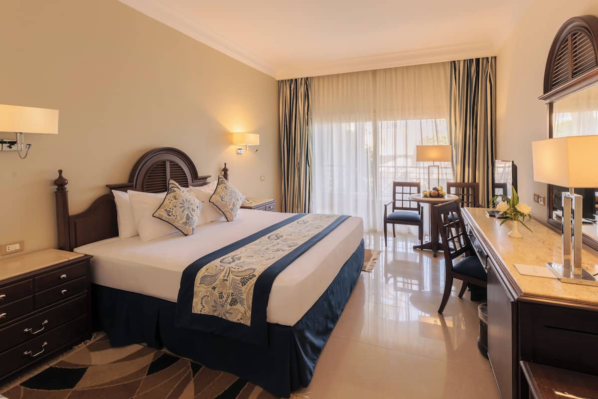Une chambre d’hôtel avec un grand lit, un bureau avec une chaise, une lampe et une fenêtre avec des rideaux.