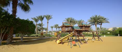 A playground with a slide and climbing structures, surrounded by palm trees and a clear sky.