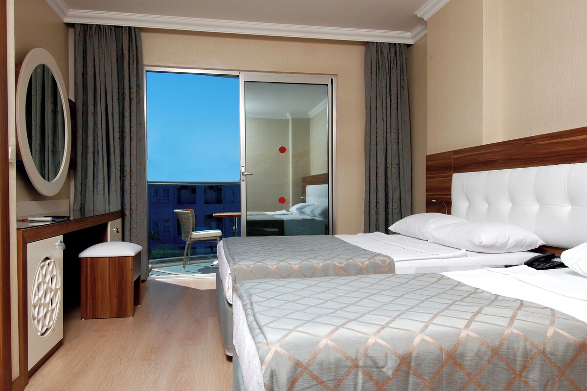 A hotel room with a bed, a desk, a chair, a mirror, and a view of the outside.