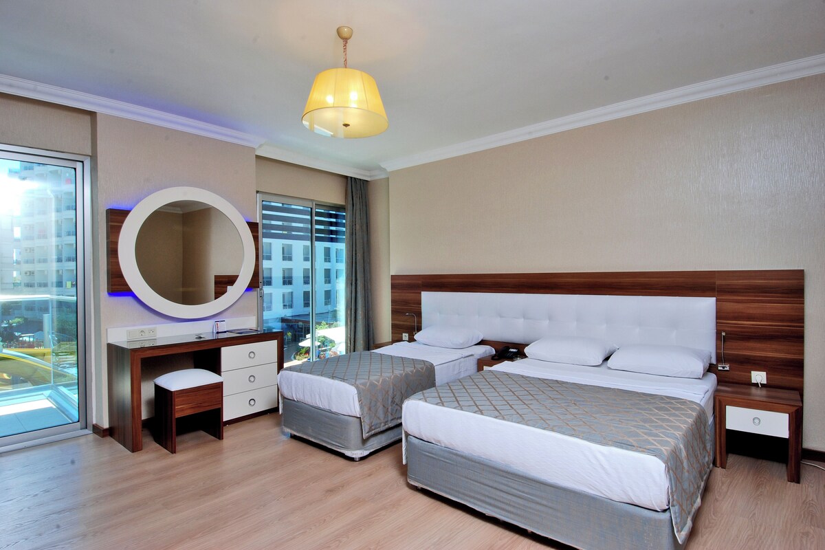 A hotel room with two beds, a desk, a mirror, and a large window.