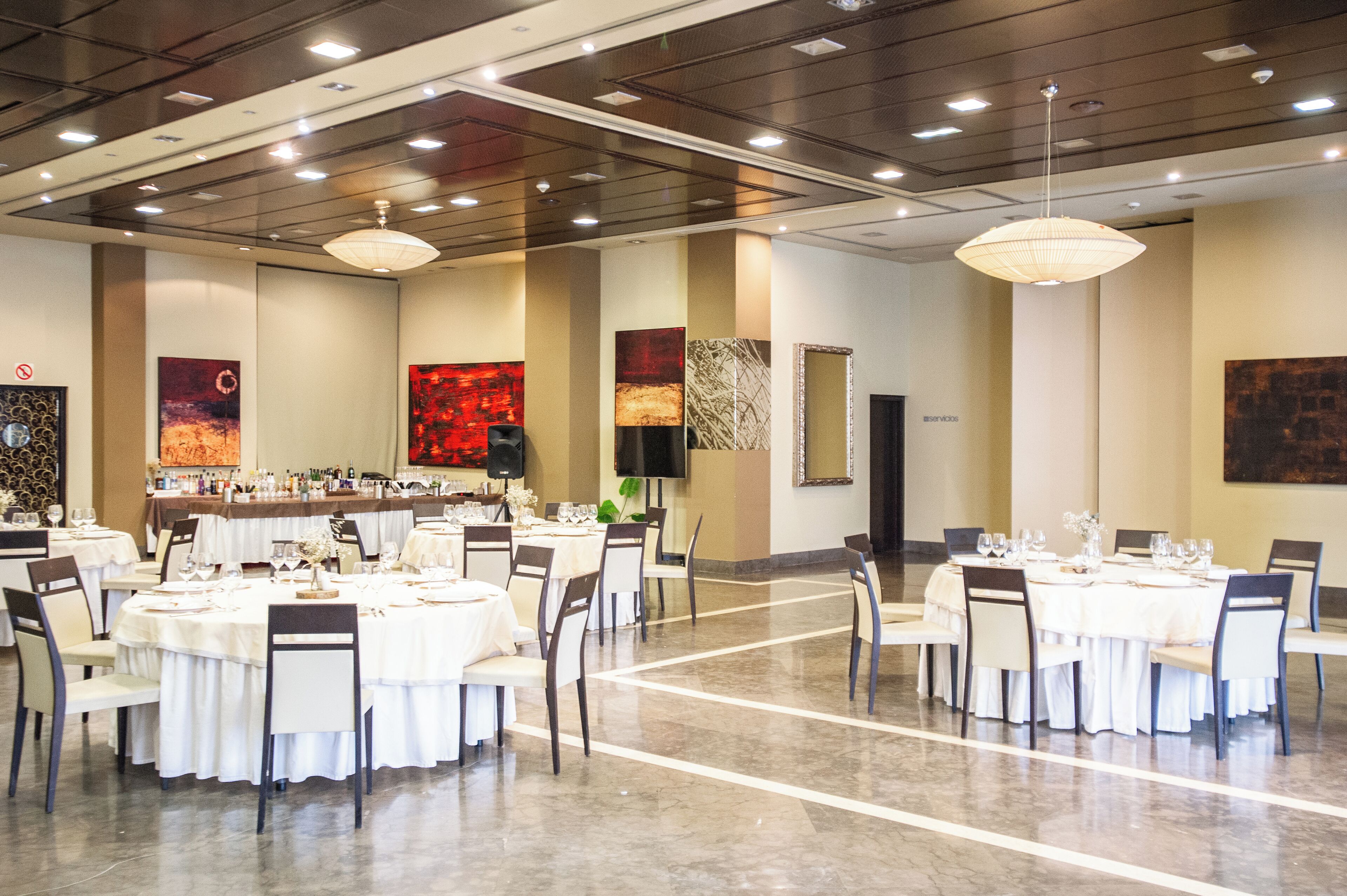 A spacious banquet hall with multiple round tables set for dining, a bar area, and modern lighting fixtures.