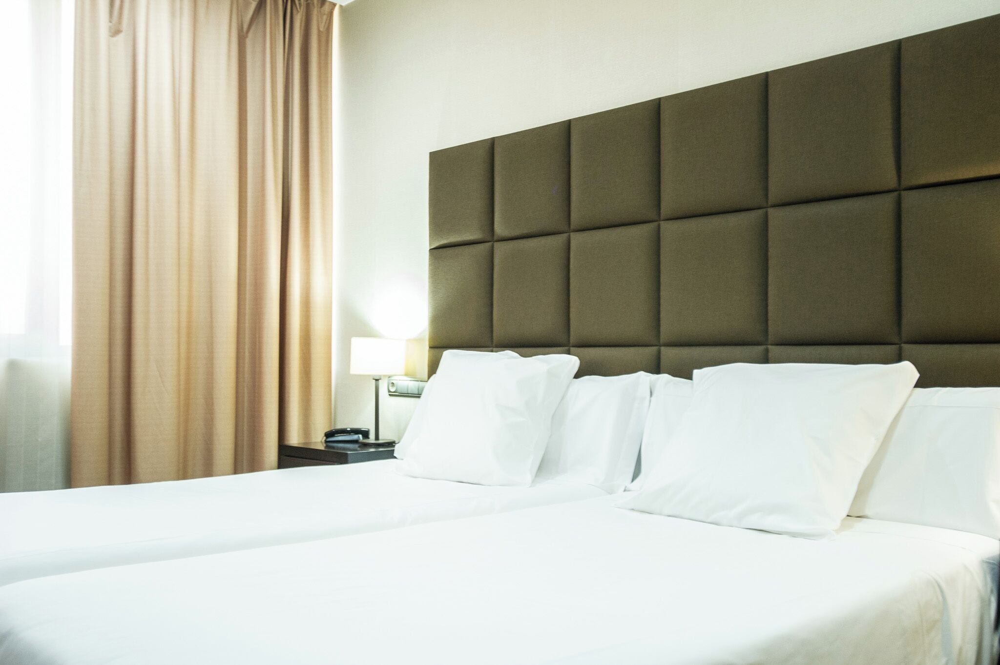 A hotel room with a large bed, a headboard with a padded design, a nightstand with a lamp, and a window with curtains.
