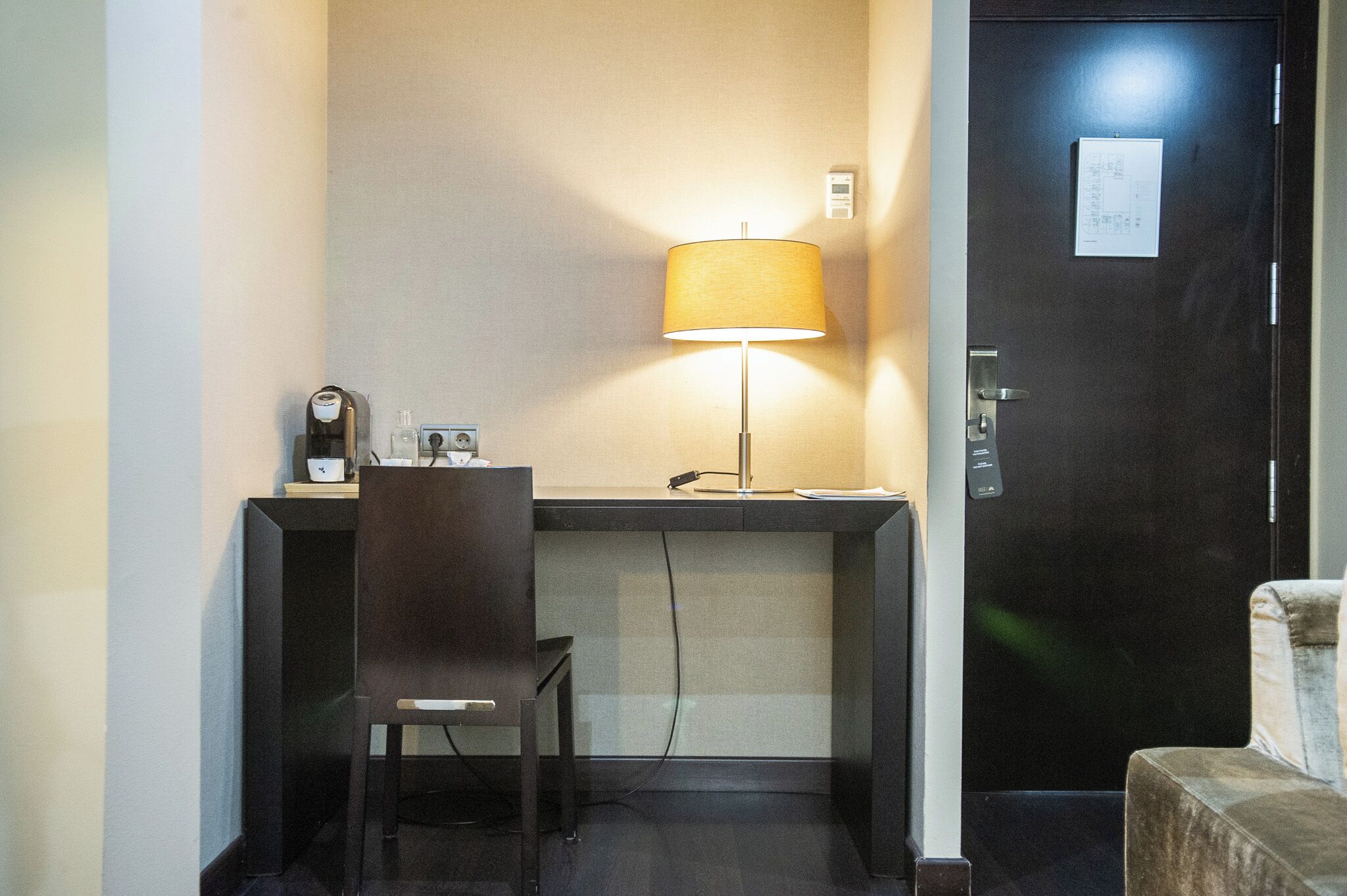 Quarto de hotel com escrivaninha, cadeira, luminária e porta com fechadura e placa.