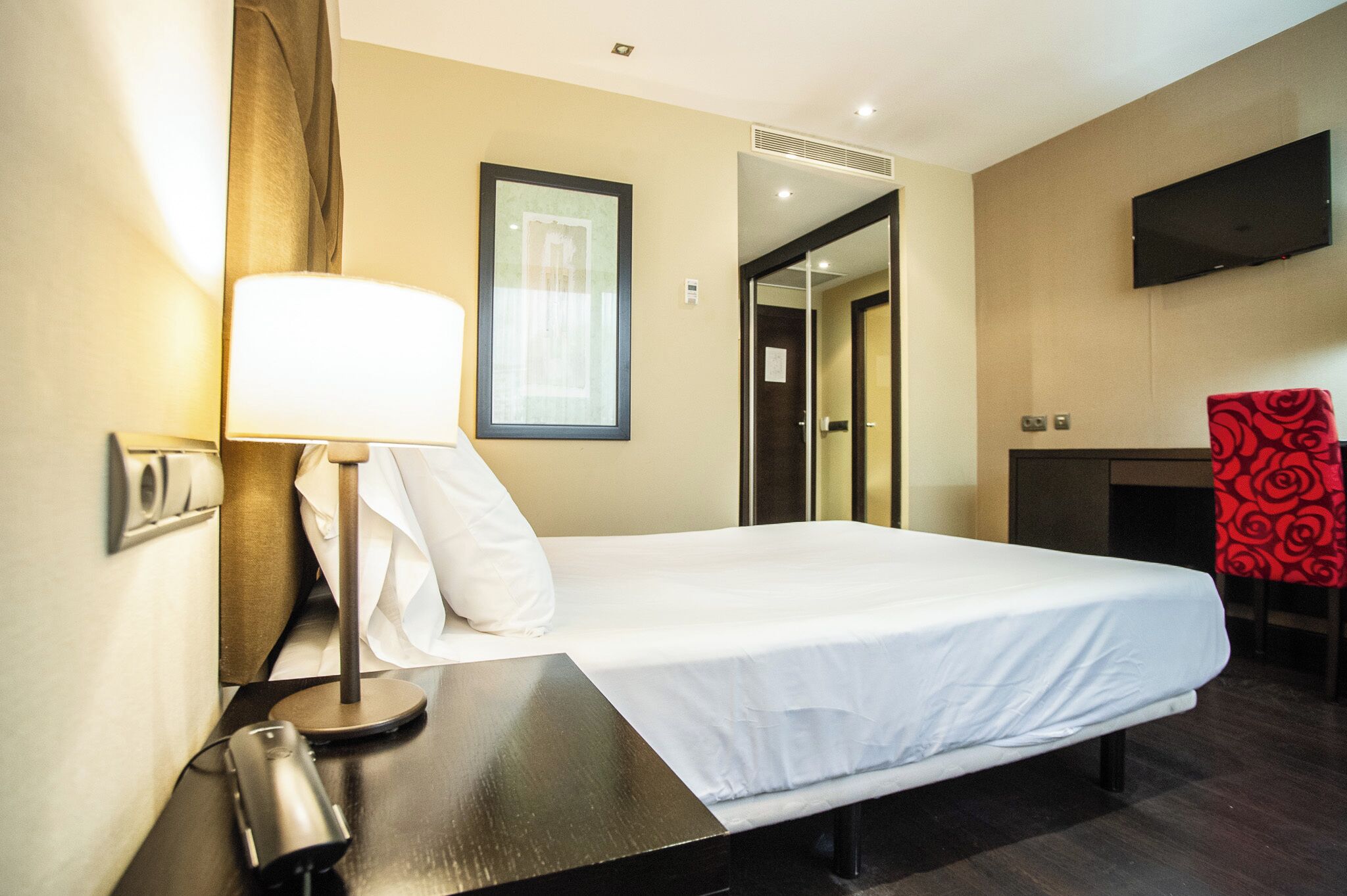 Ein Hotelzimmer mit Bett, Schreibtisch mit Lampe, Fernseher und einem roten Sessel.