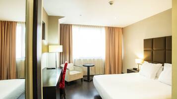 Habitación de hotel con una cama grande, un escritorio con silla, una mesita pequeña y una mesita de noche con lámpara.