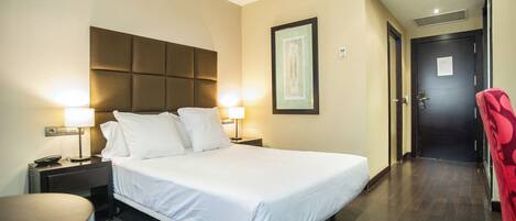 A hotel room with a large bed, bedside lamps, a framed picture on the wall, and a door in the background.