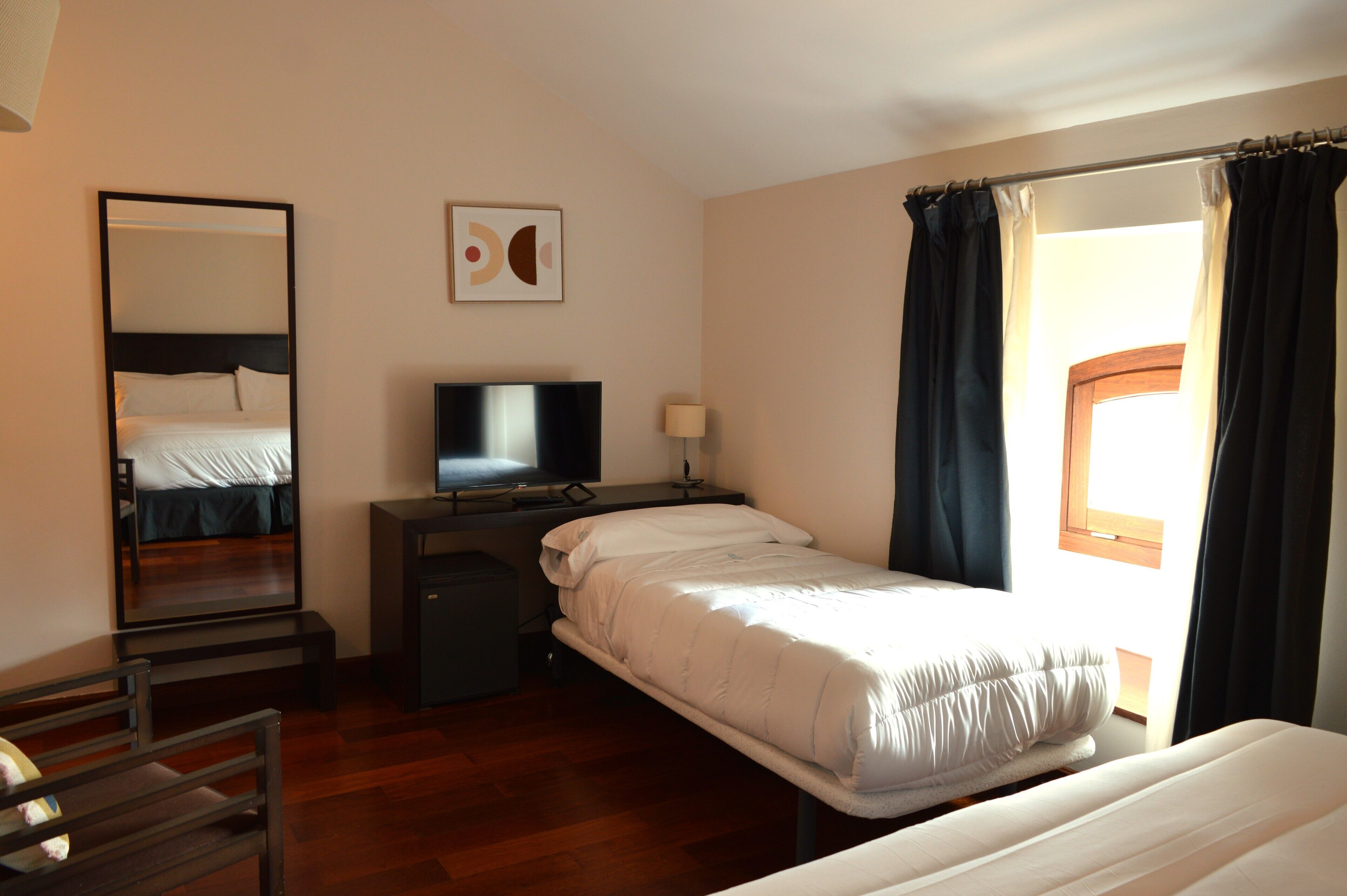 Une chambre d’hôtel avec un lit, un bureau avec une télévision, un miroir et une fenêtre avec des rideaux.