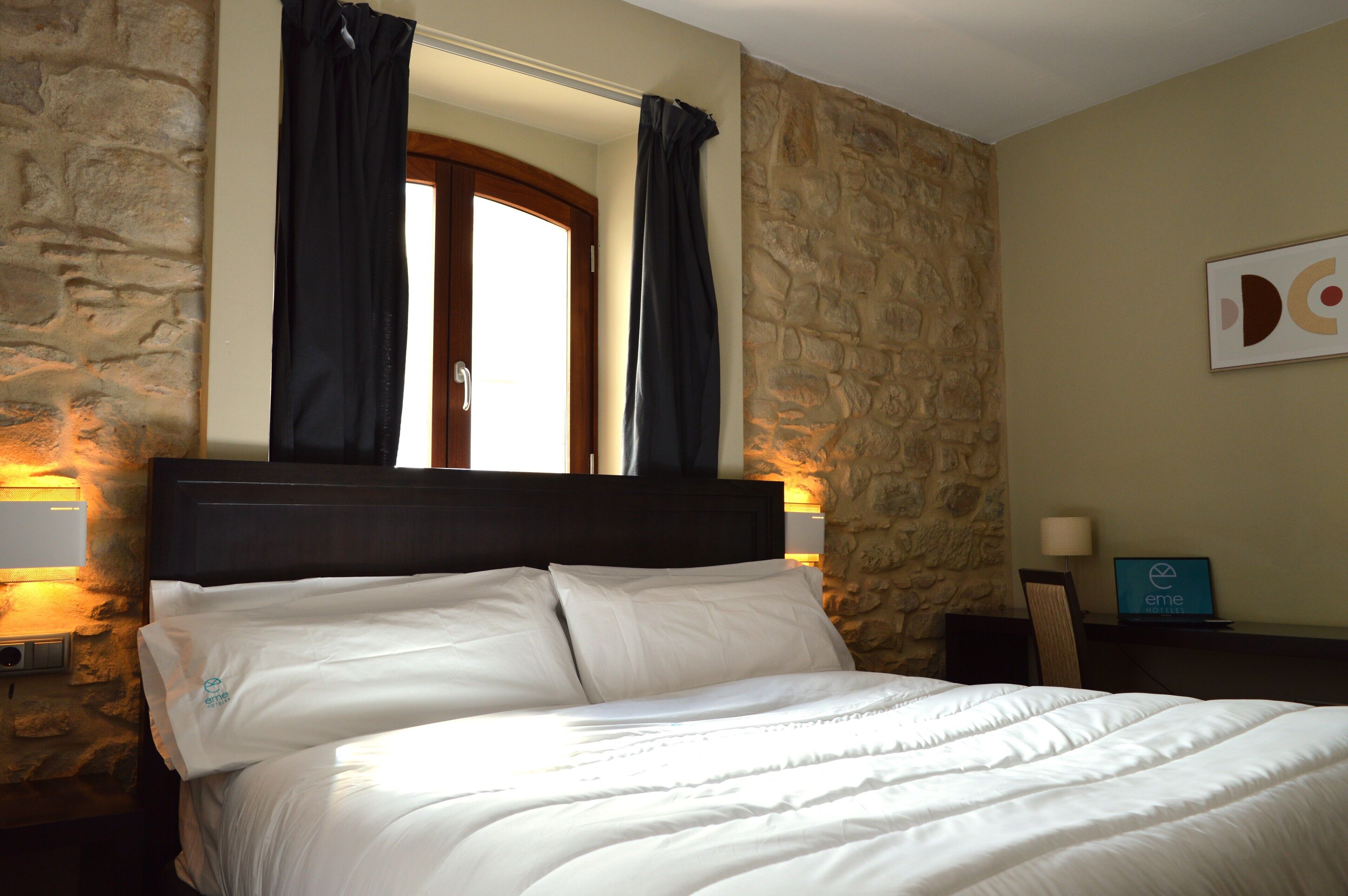 A bed with white bedding, a stone wall, a wooden headboard, and a window with curtains.