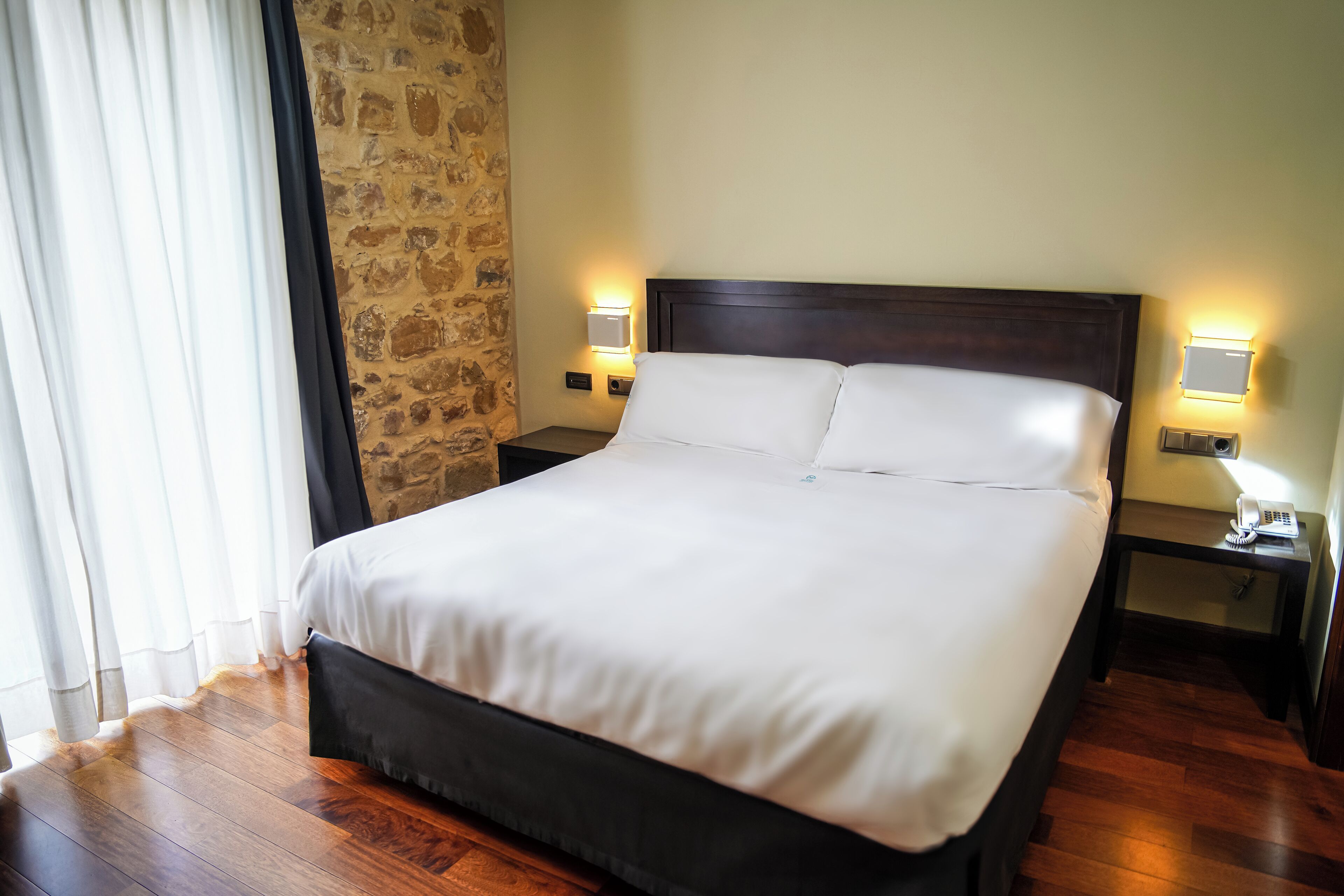 A neatly made bed in a hotel room with a stone wall, wooden flooring, and bedside lamps.