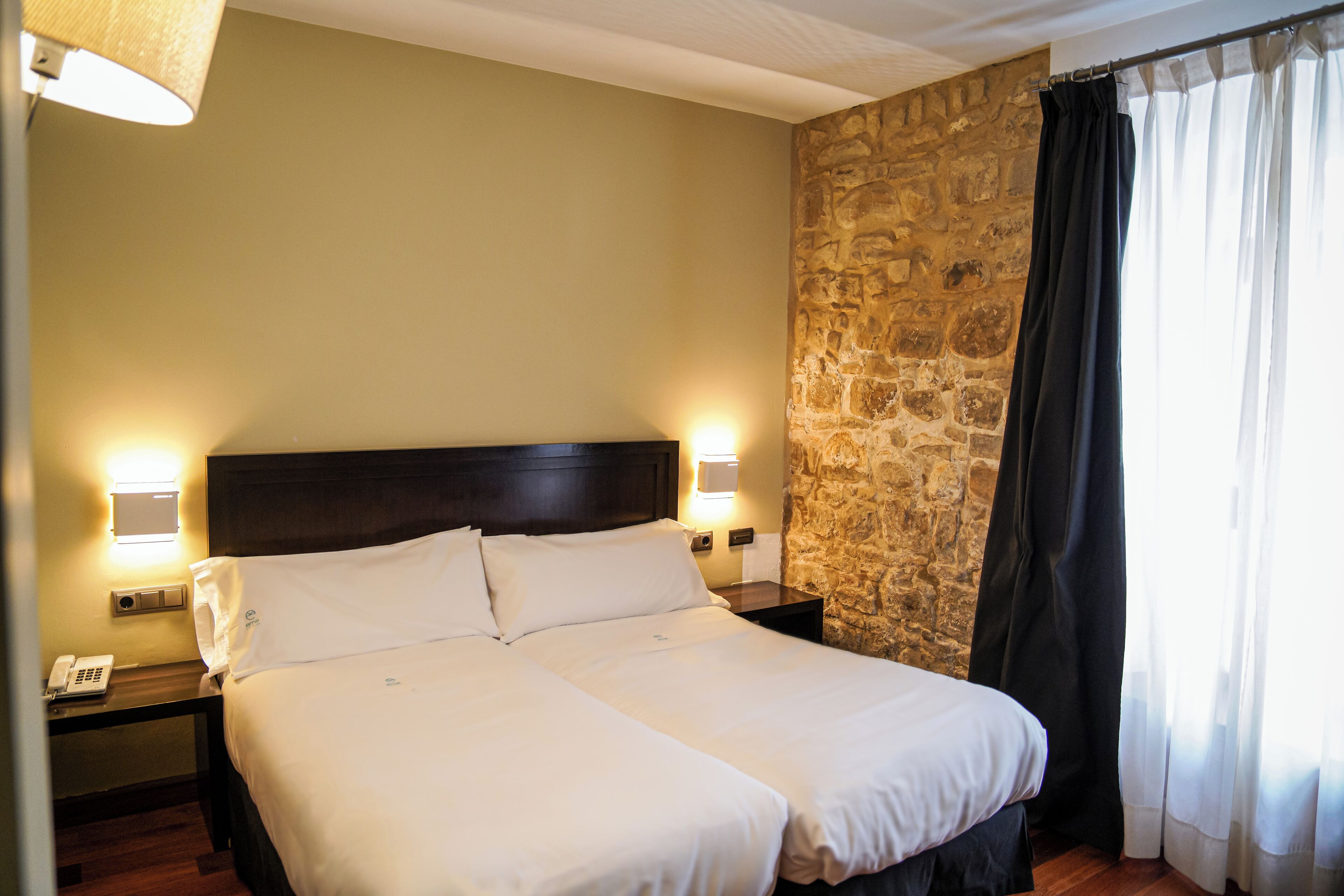 A double bed with white linens, two bedside tables with lamps, a stone wall, and a window with black curtains.