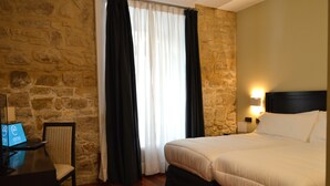 A hotel room with a stone wall, a bed with white bedding, a desk with a laptop, and a chair. - Hotel Baeza Monumental by eme Hoteles (Baeza)