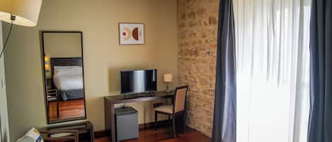 A hotel room with a wooden floor, a desk with a television, a chair, a small cabinet, a bed, a mirror, and a window with curtains.