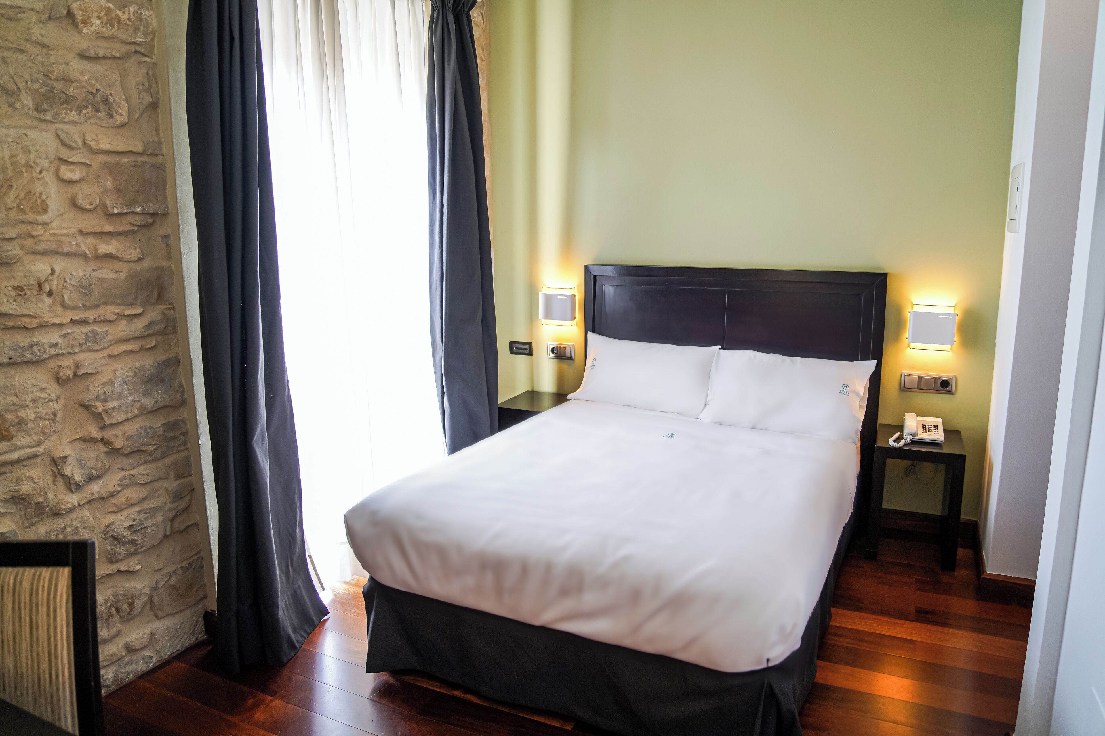 A neatly made bed with white linens, two bedside lamps, a nightstand with a phone, and a stone wall.