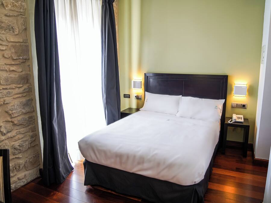 A neatly made bed with white linens, two bedside lamps, a nightstand with a phone, and a stone wall.