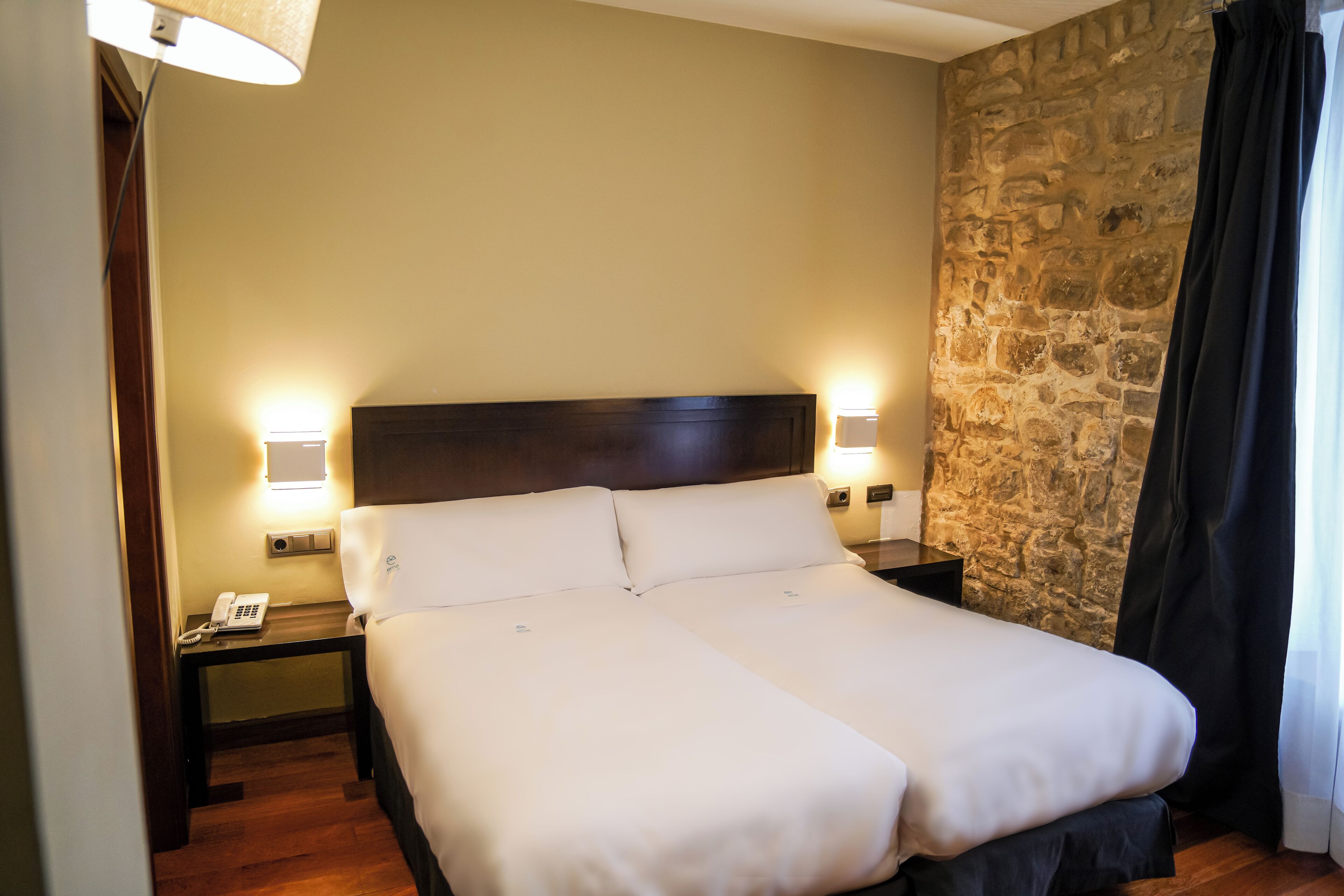 Un lit double avec du linge de lit blanc, dans une pièce aux murs en pierre et dotée d’une tête de lit en bois. On y trouve deux tables de chevet, chacune avec une lampe, et une table porte un téléphone.