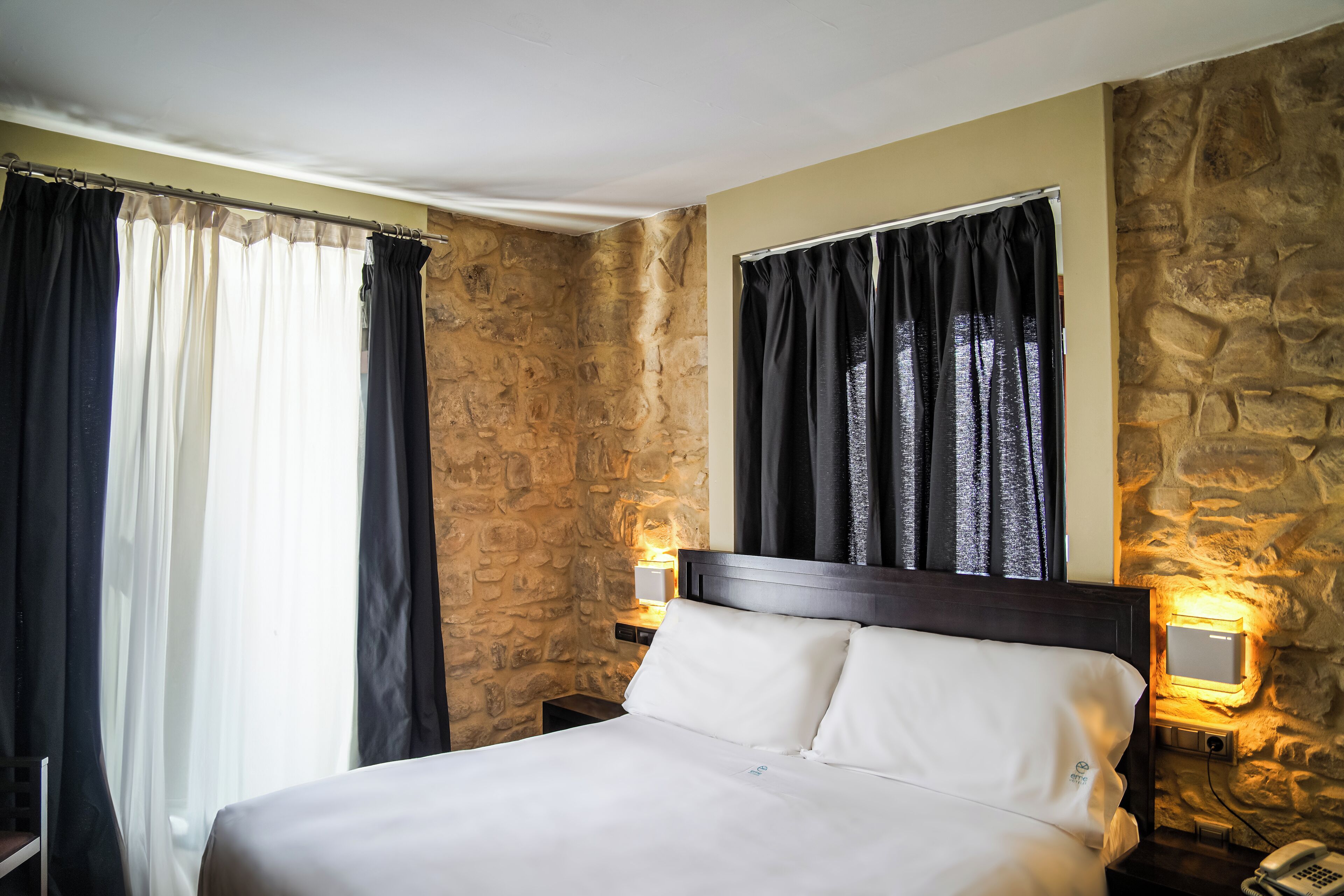 Camera d'albergo con parete in pietra, un letto con lenzuola bianche, un comodino con una lampada e un telefono.