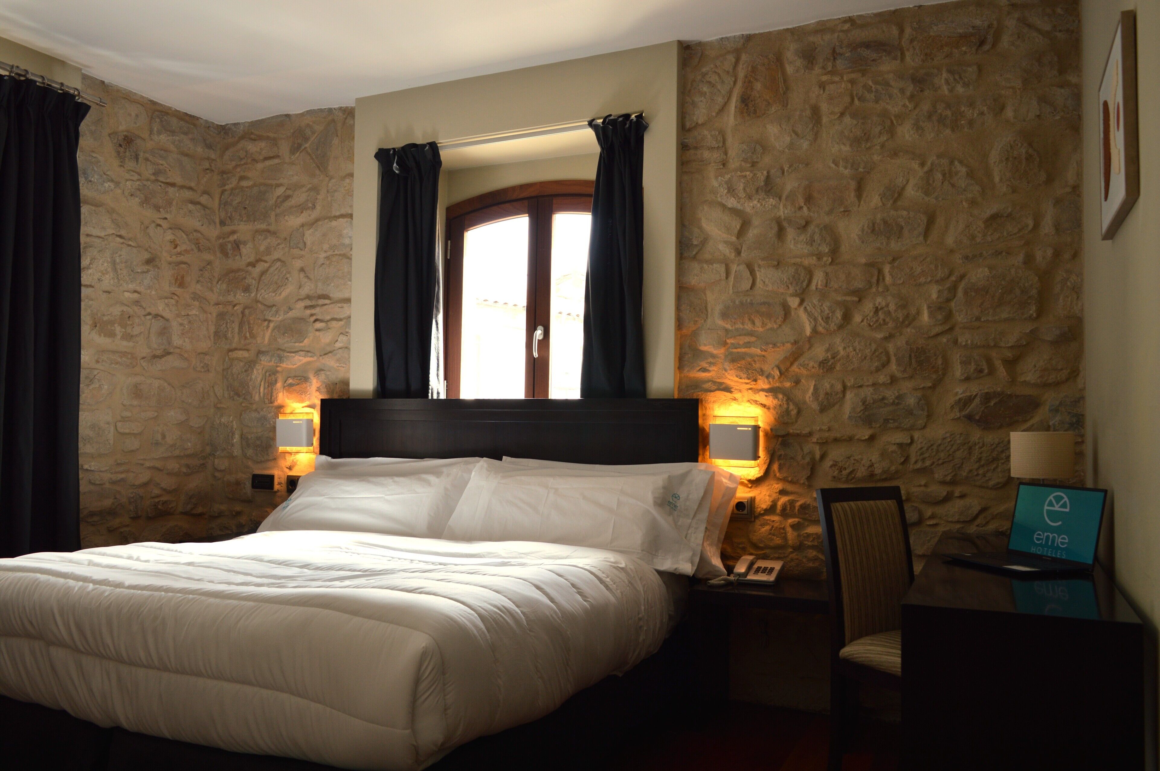 A hotel room with a stone wall, a bed with white bedding, a wooden headboard, two bedside tables with lamps, a chair, and a desk with a laptop displaying the hotel logo.