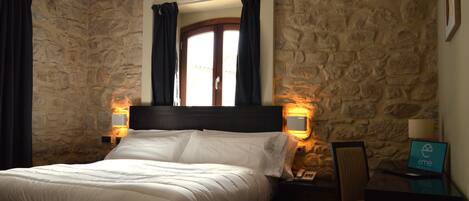 Habitación de hotel con pared de piedra, cama con ropa blanca, cabecero de madera, dos mesitas de noche con lámparas, una silla y un escritorio con una laptop mostrando el logotipo del hotel.