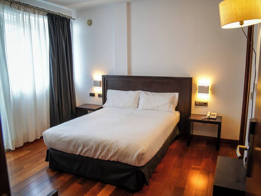 Una cama bien tendida con sábanas blancas en una habitación de hotel con suelo de madera, dos mesitas de noche con lámparas y una ventana con cortinas.