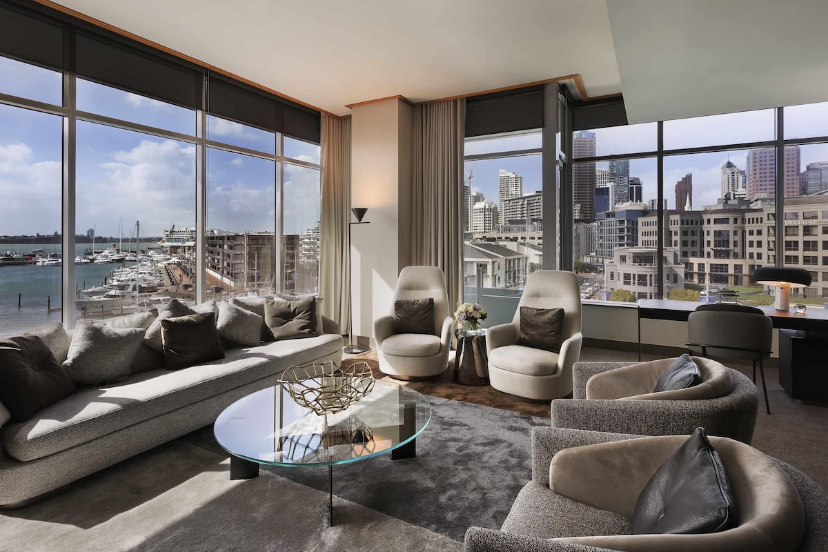 A modern living room with a sofa, armchairs, a glass coffee table, and a view of a marina and cityscape.