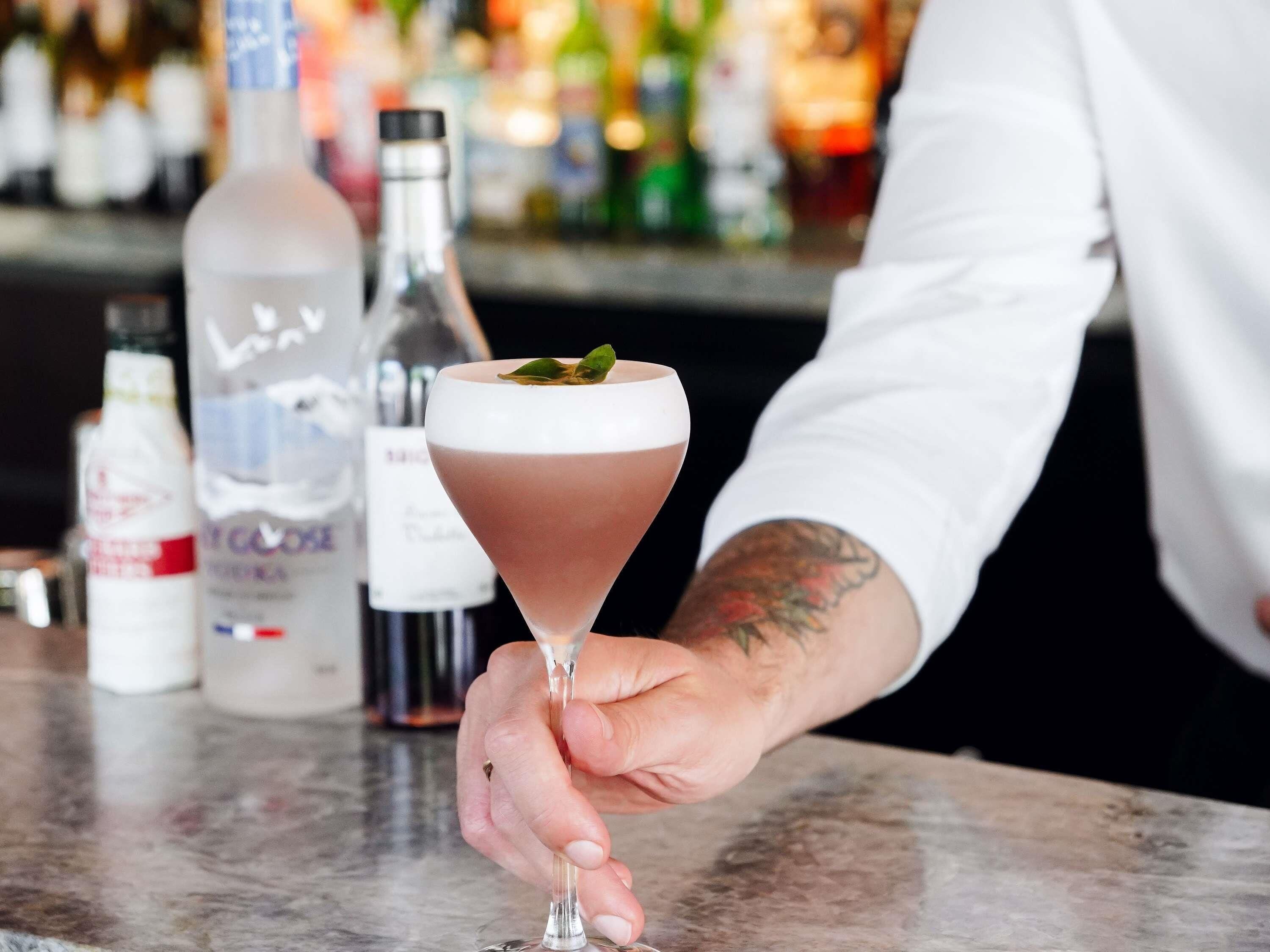 A person holding a cocktail glass with a layered drink and a garnish, with bottles of alcohol in the background.