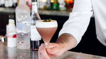 A person holding a cocktail glass with a layered drink and a garnish, with bottles of alcohol in the background.