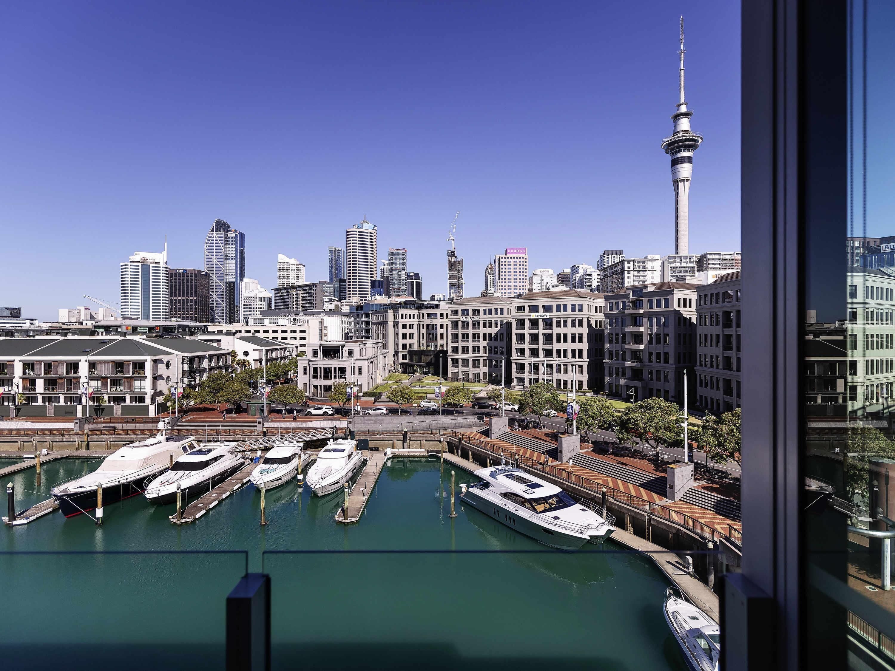 A marina with several yachts docked, a city skyline with modern buildings, and a prominent tower.