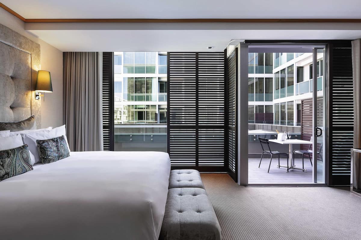 A modern hotel room with a large bed, a seating area, and a balcony with a table and chairs.