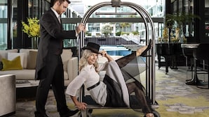 A man in a suit assisting a woman in a wheelchair through a modern lobby with a pool view, a chandelier, and a patterned floor. - Sofitel Auckland Viaduct Harbour (Auckland)